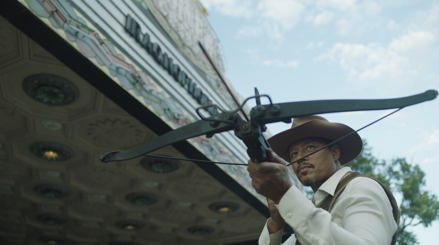 Terrence Howard holds a crossbow in front of a movie marquee while wearing a cowboy hat in Showdown at the Grand.