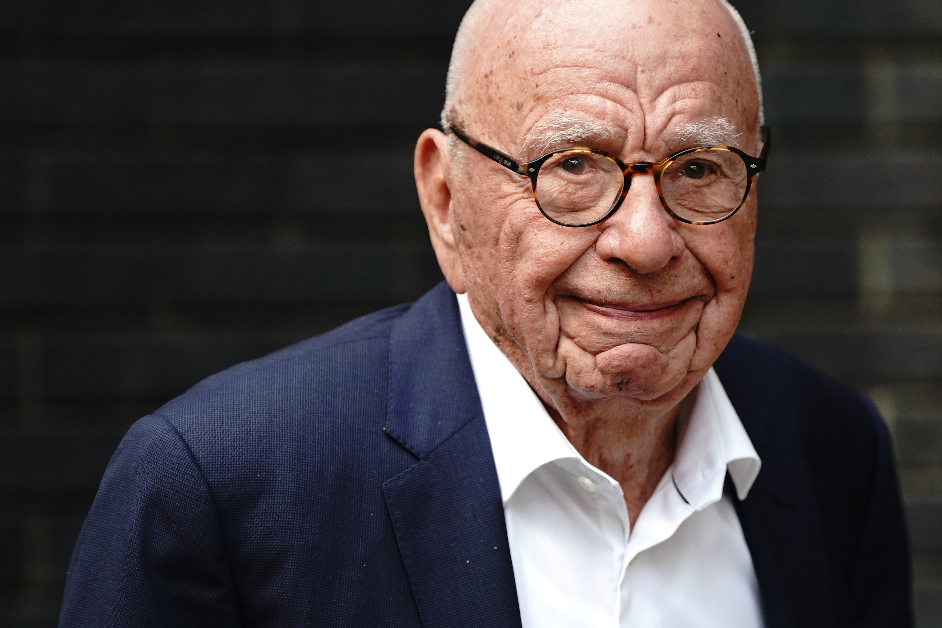 Rupert Murdoch with a slight smile, wearing a blue suit jacket and white shirt open at the throat.