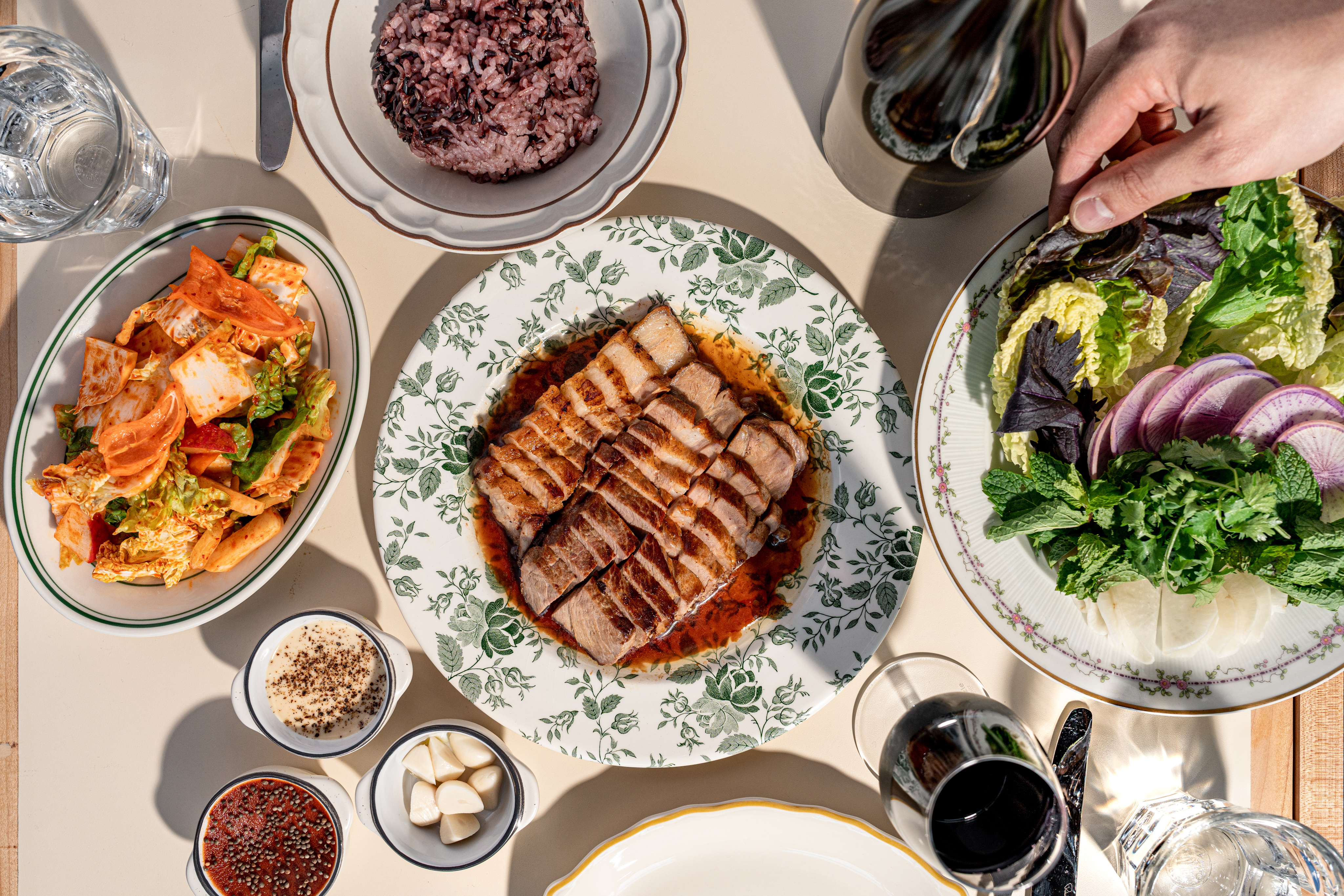 A hand places ssam lettuces at a table with pork coppa, banchan, kimchi, and more.