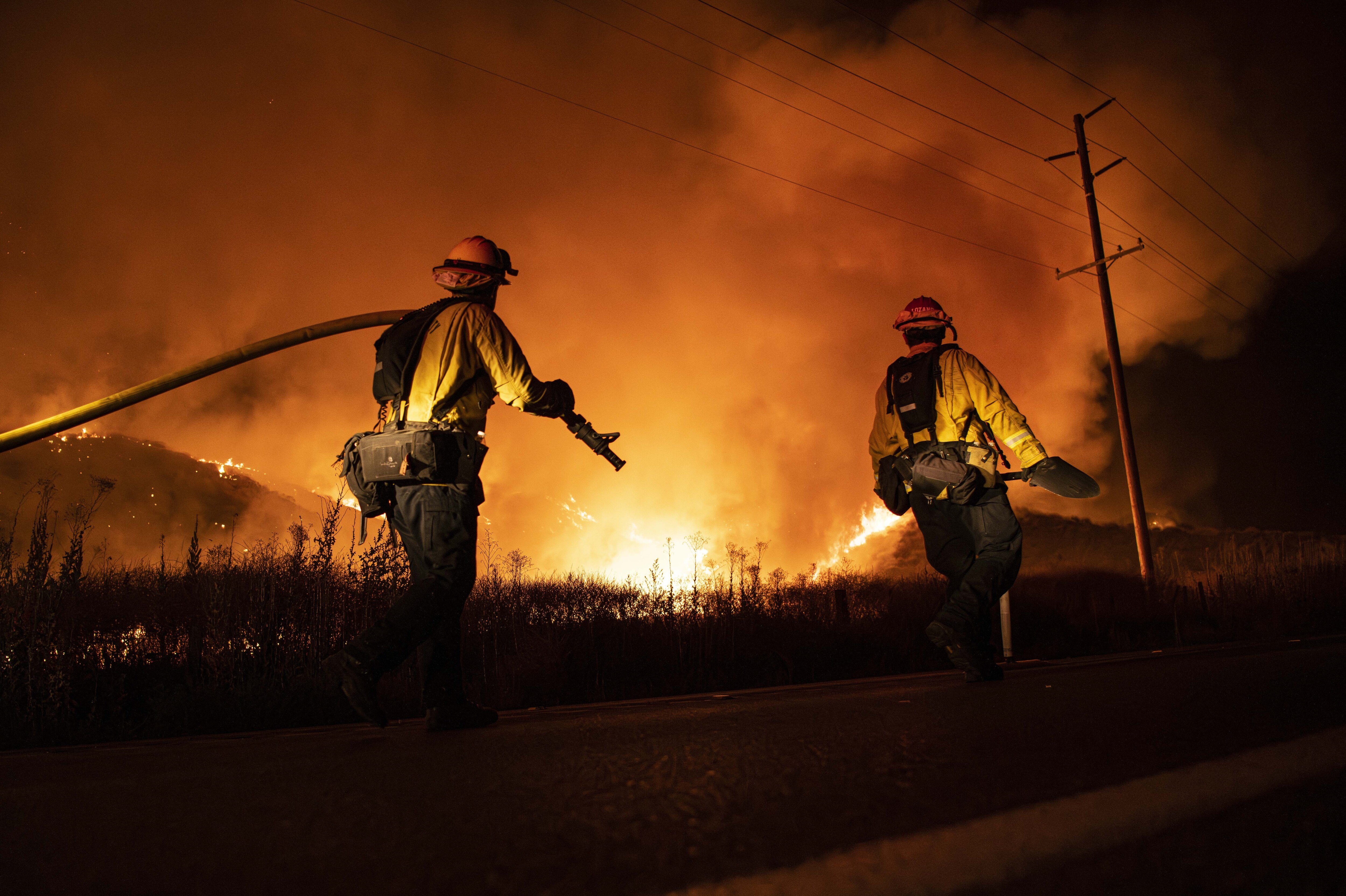 Two firefighters, one carrying a hose, are illuminated by a wildfire burning in the background.