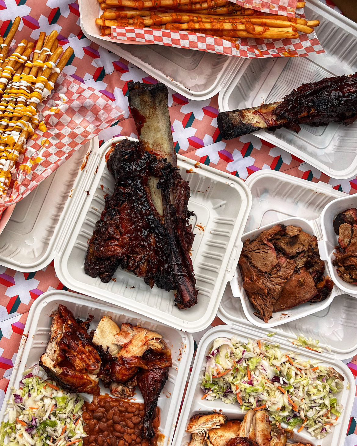 Several takeout containers of barbecue ribs and other barbecue-style dishes set on top of a red and white-checkered tablecloth.
