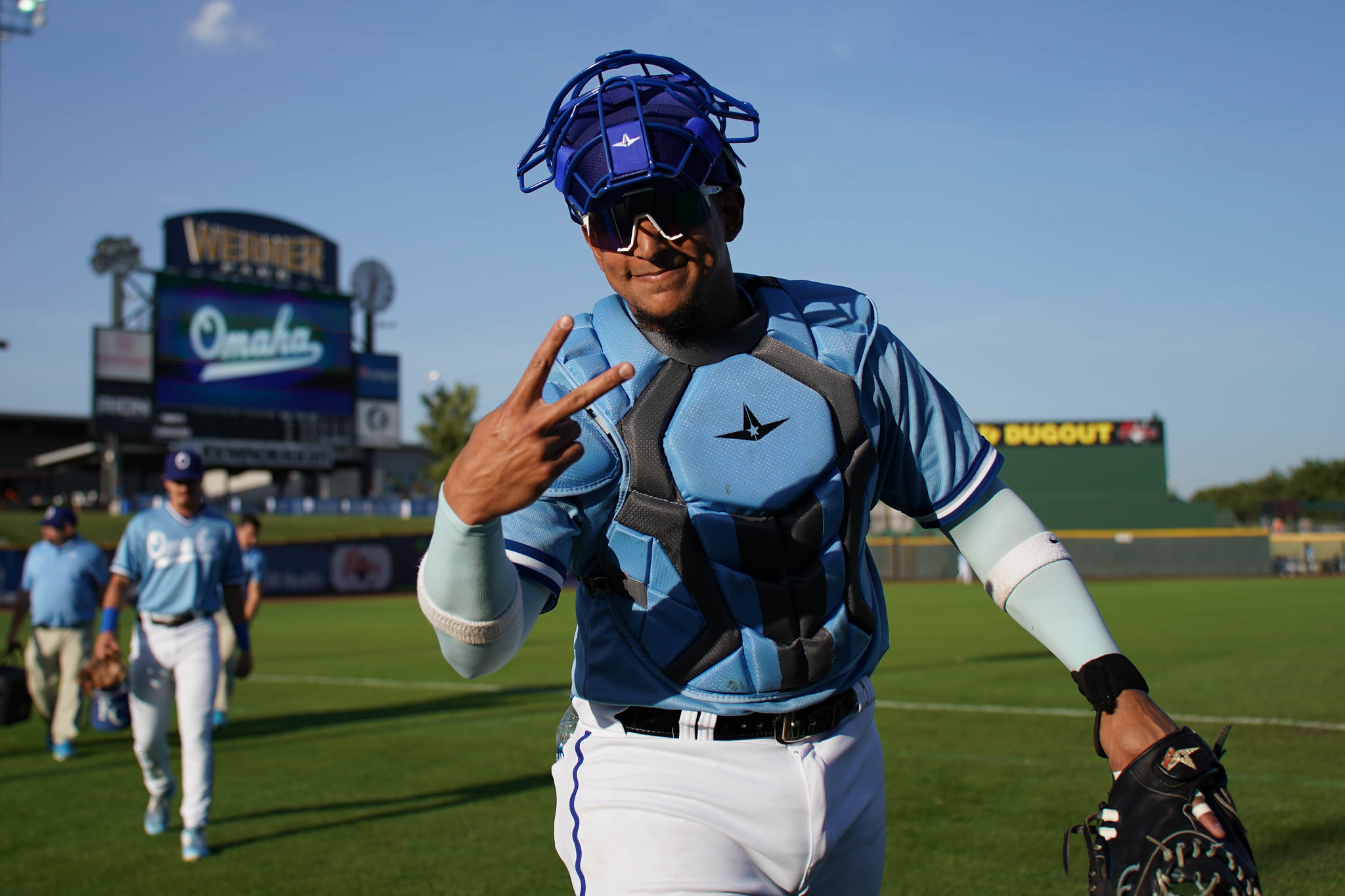 A catcher in light blue and gray chest protector gives a peace sign as he walks on a baseball field.