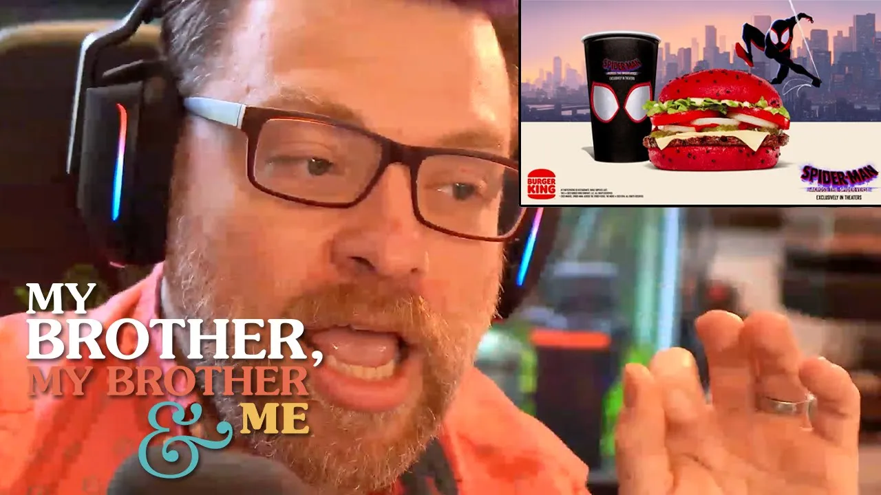 A photograph of Travis McElroy, close to the camera. He is wearing glasses and headphones, and he has his left hand raised, just visible in frame. In the upper right hand corner is an ad for the Burger King Spider Man Whopper, in the lower left hand corner is the MBMBaM logo.