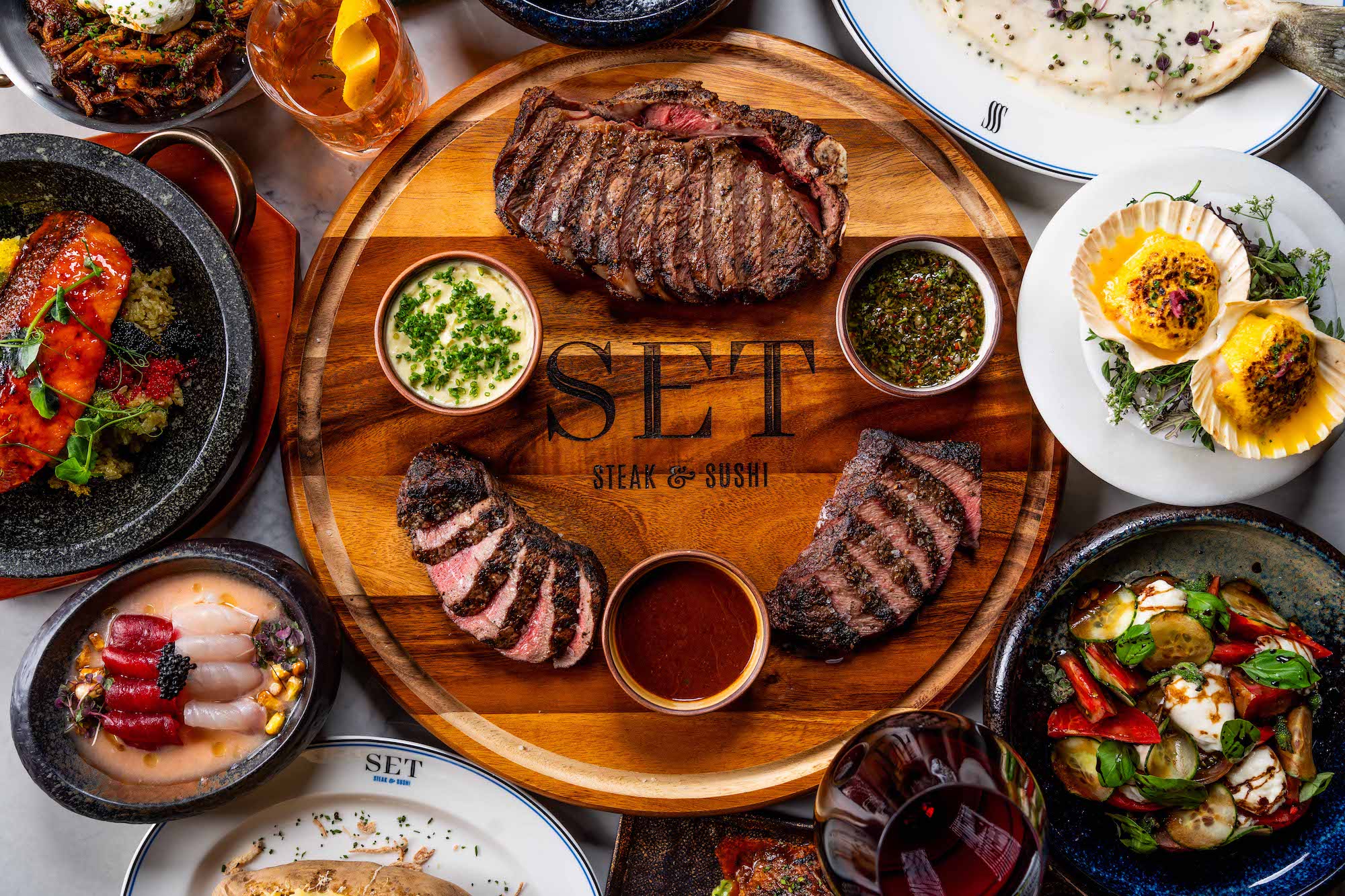 An overhead shot of a grilled steak sliced onto a dark round cutting board and surrounded by other dishes like scallops and vegetables.