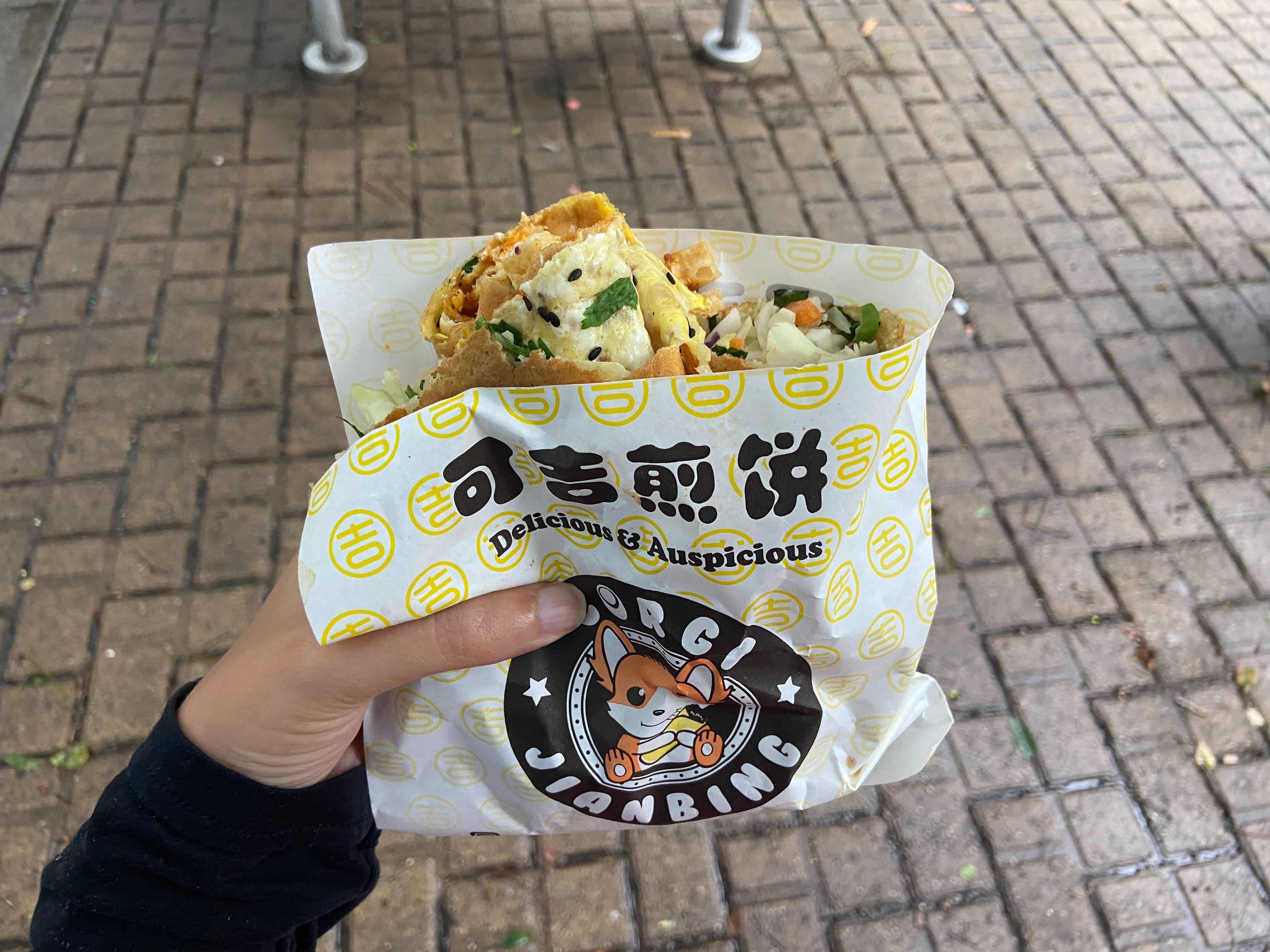 A hand holds up a jianbing in wrapping with a corgi cartoon character.