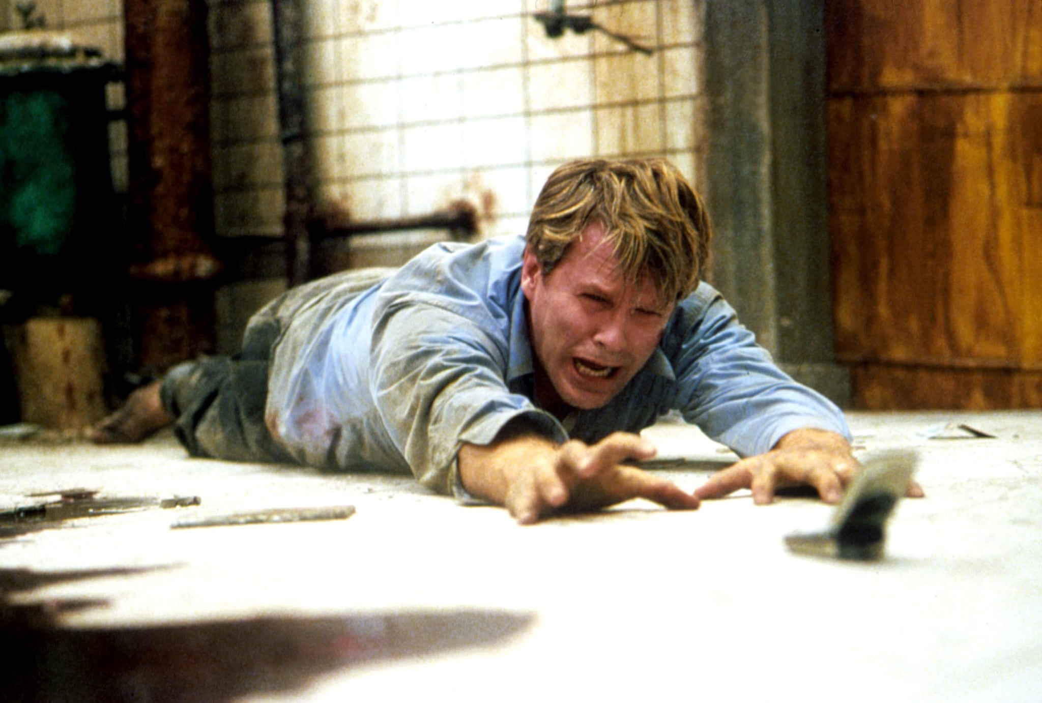 Cary Elwes, rumpled and covered in filth, lies on a dirty basement floor and weeps as he tries to grab a cell phone lying on the floor just out of reach in the original 2004 Saw