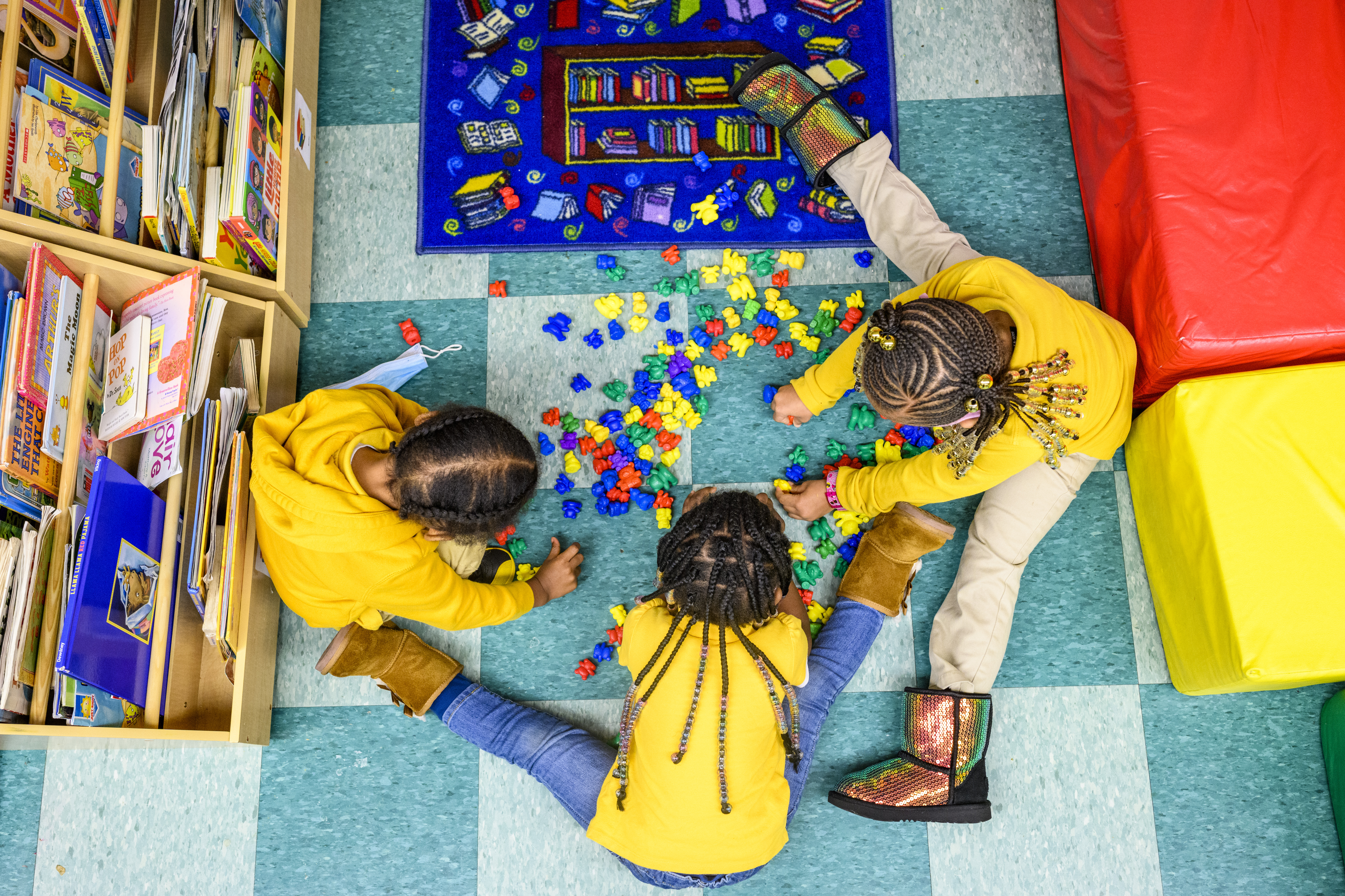 Seen from above, three children in yellow shirts play with brightly colored puzzle pieces on a tiled floor. 