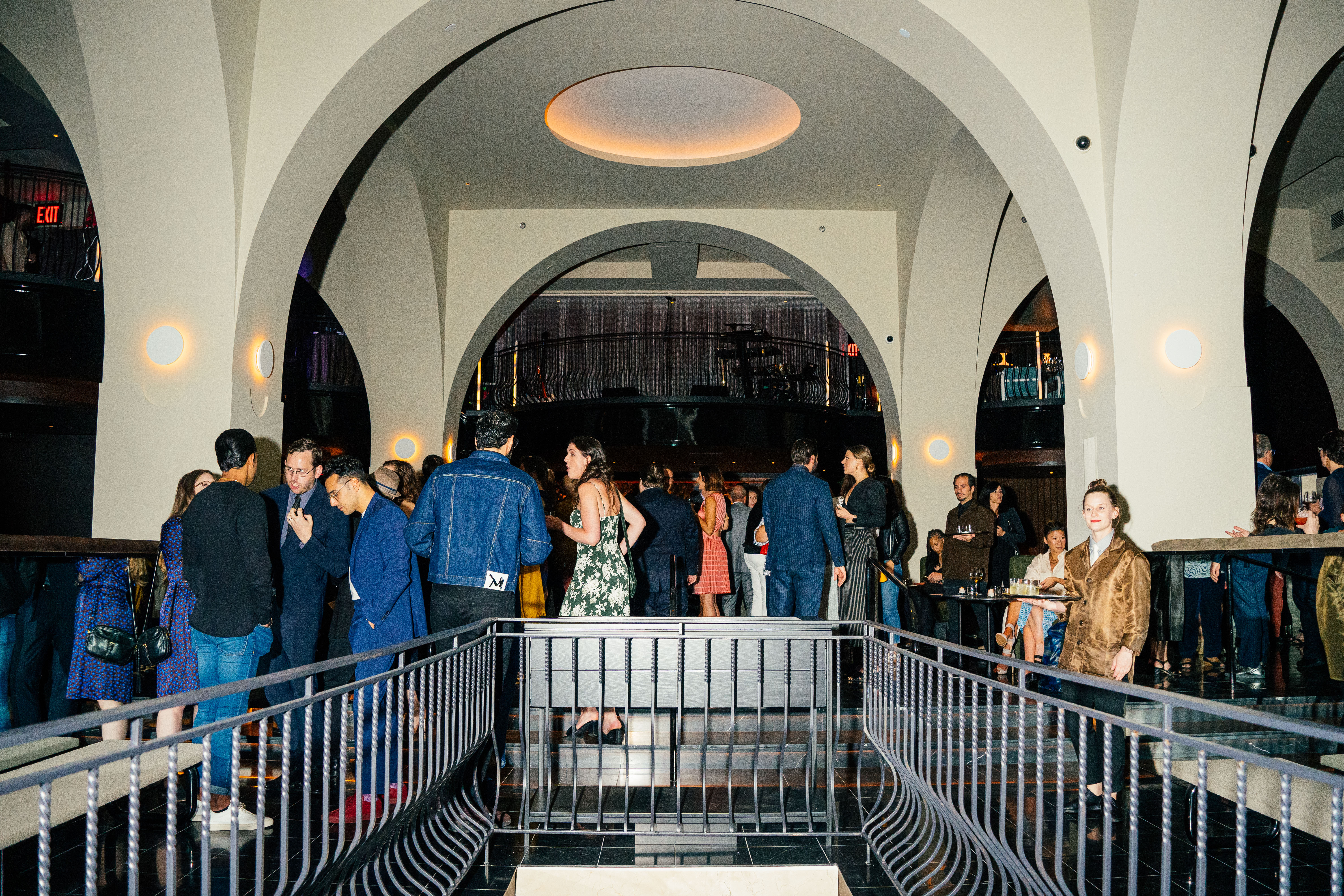 Well-dressed customers mingle in an arched dining room with a stage in the background.