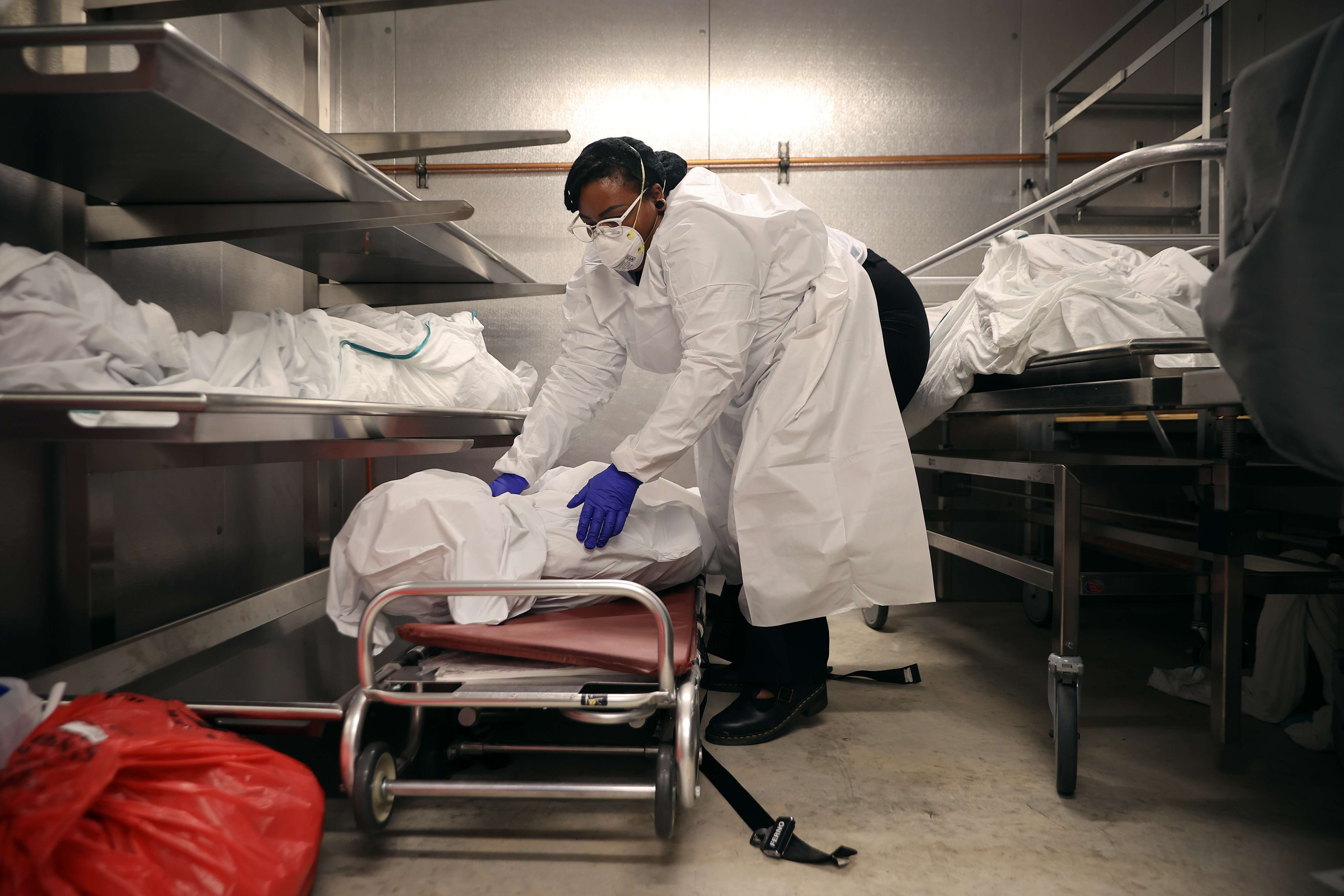 A worker wearing a mask, surgical gown, and gloves places a body wrapped in a sheet onto a stretcher inside a morgue.