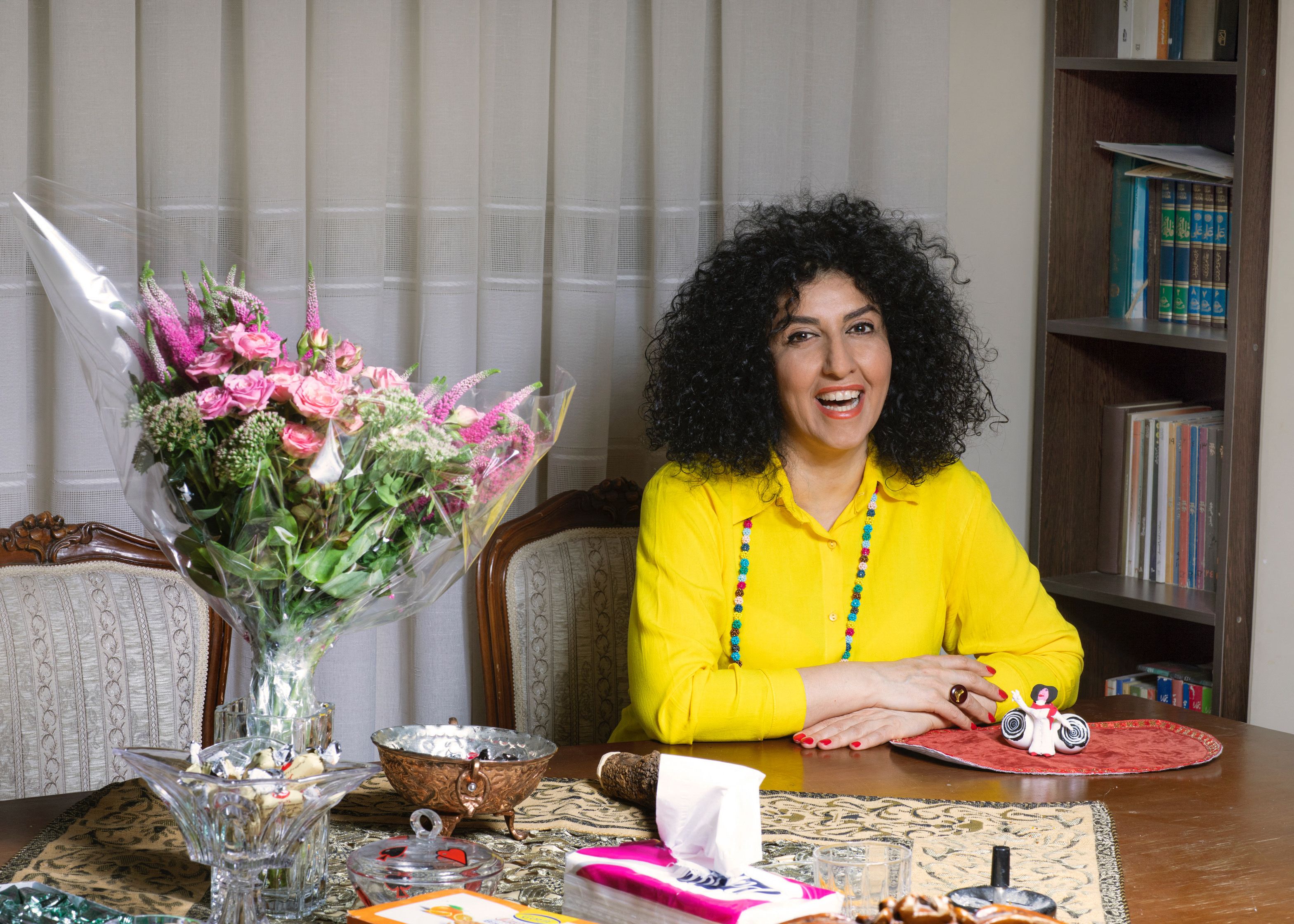 Narges Mohammadi sits at a wooden table beside a bouquet of pink flowers. She wears a bright yellow blouse and smiles broadly. 