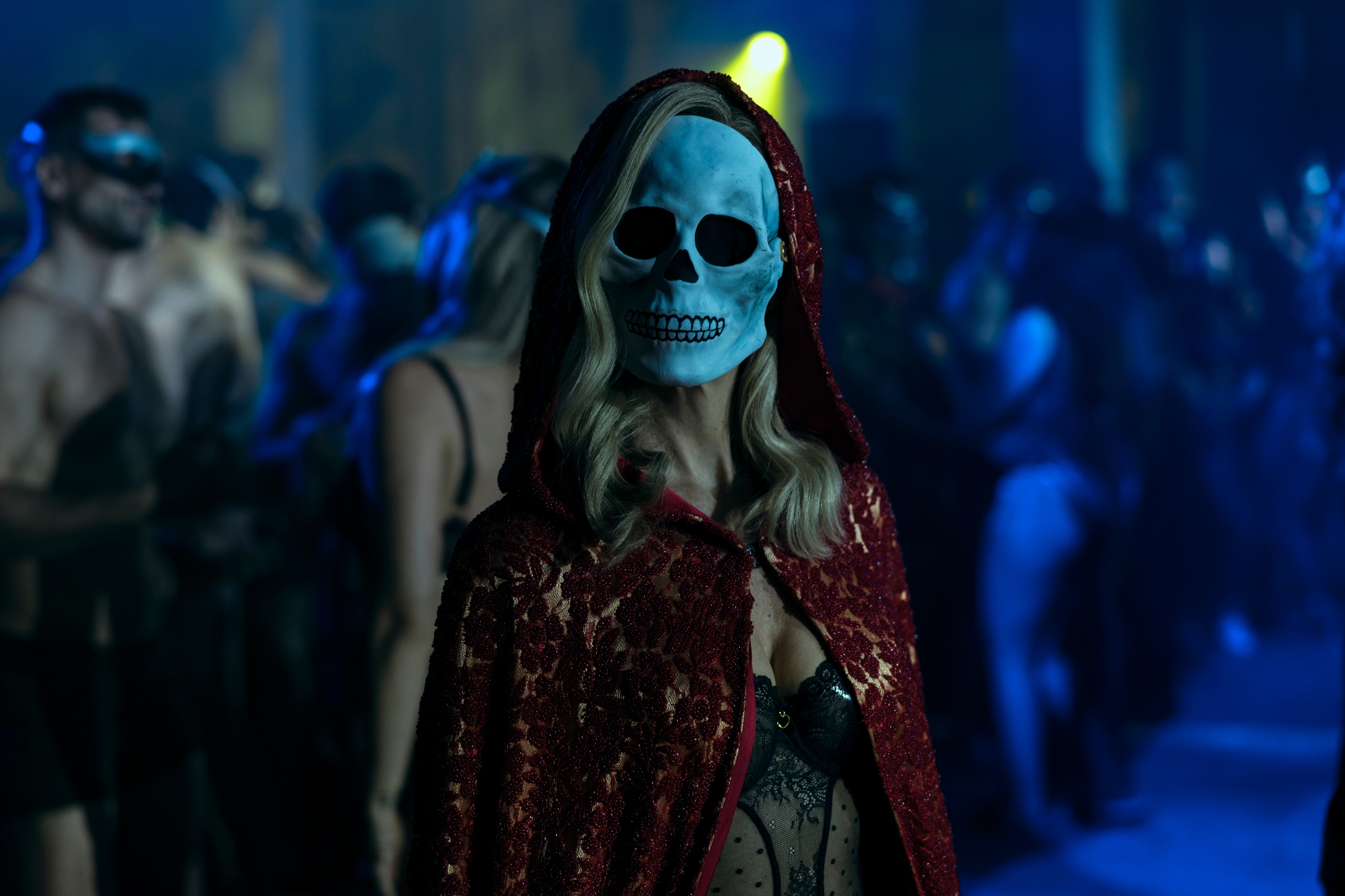 A woman wearing a red, lacy hooded cape, whose actual face is hidden by a skull mask, appears to stare directly at the camera. 