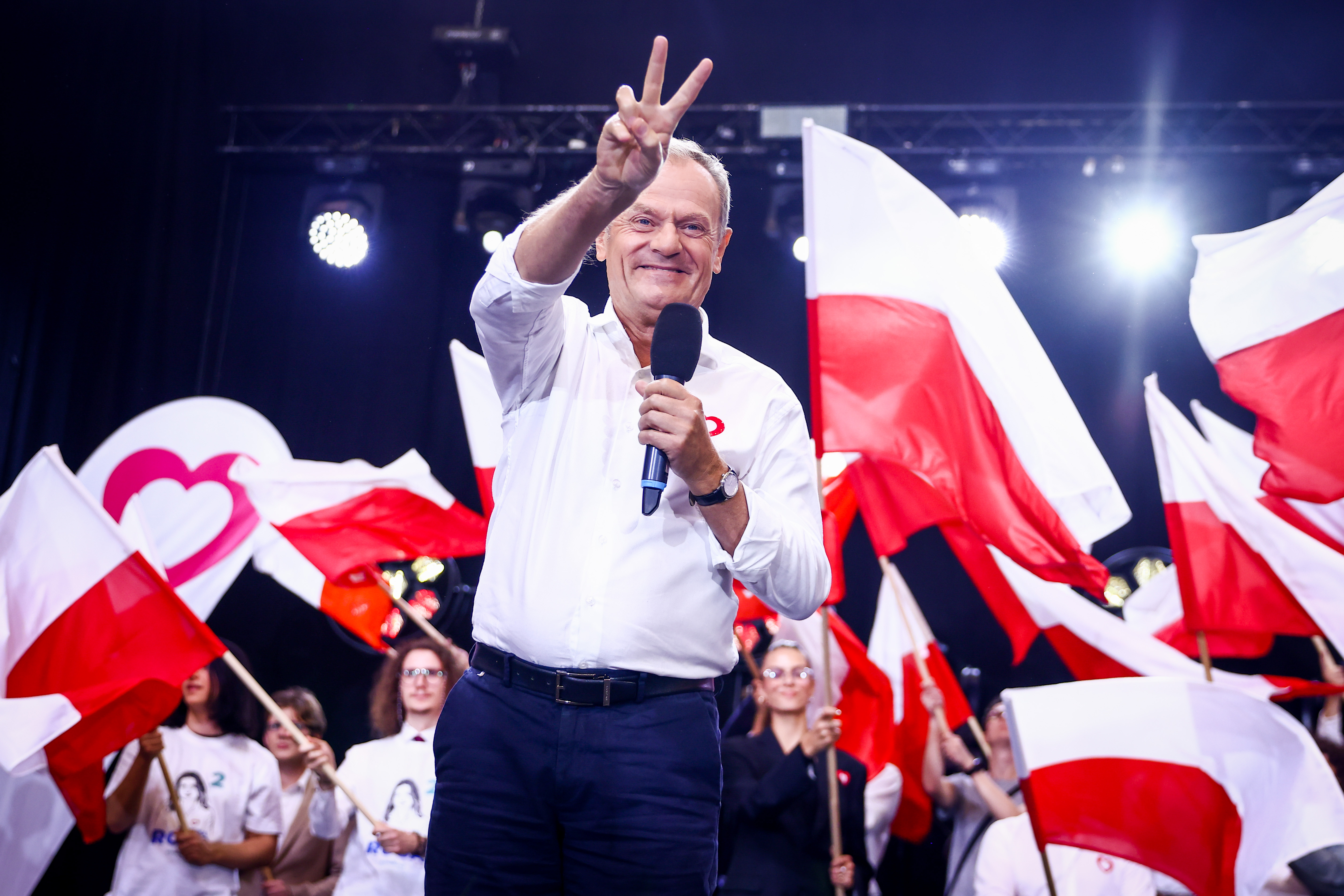 Donald Tusk, amid waving red-and-white flags, holds a microphone in one hand and makes a V-sign with the other.