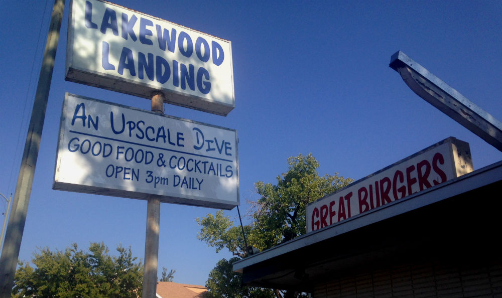 The exterior of a dive bar called Lakewood Landing. A blue and white sign identifies it as “an upscale dive” with “good food and cocktails.”