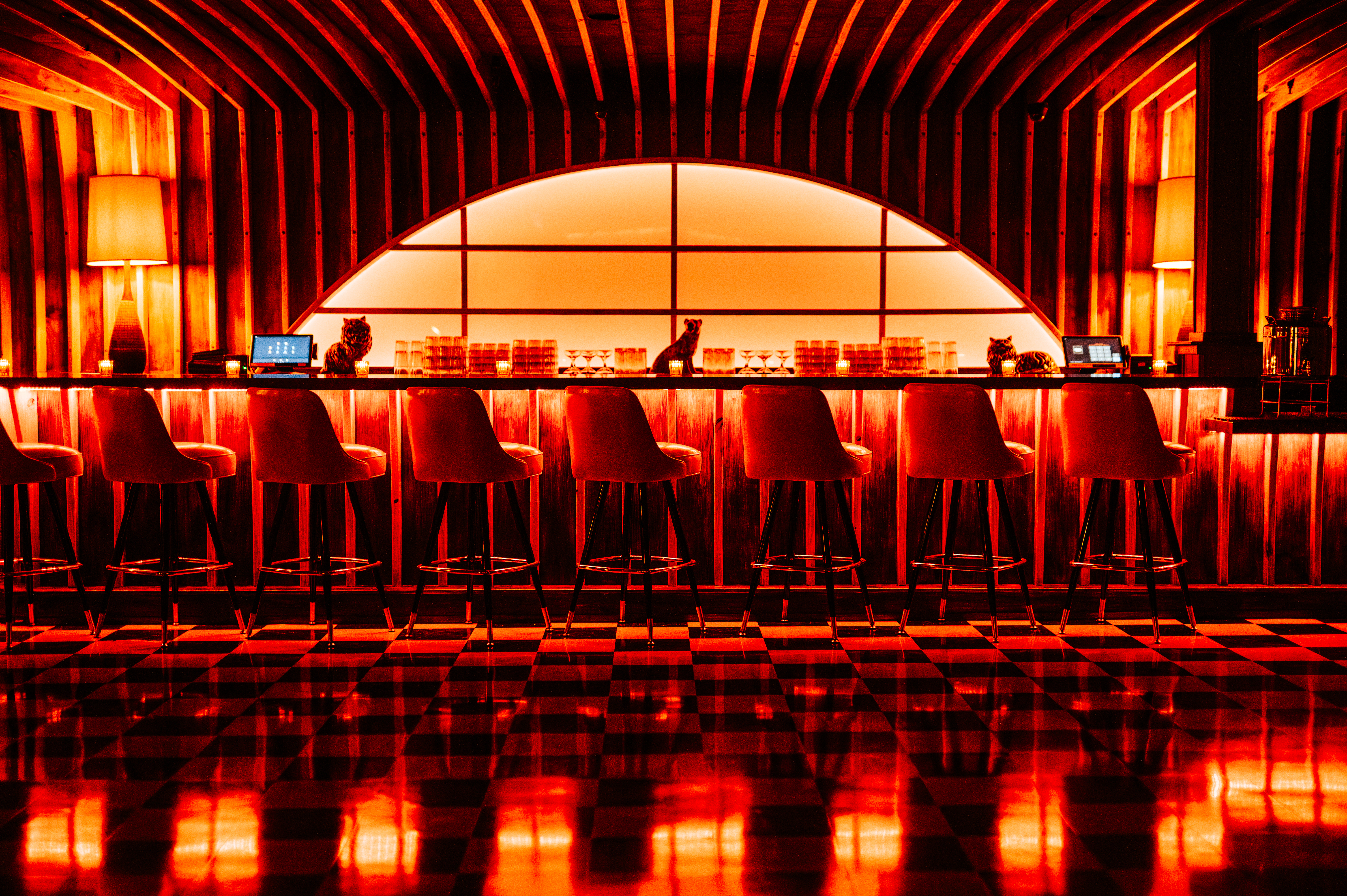 A red-hued, dimly lit stretch of bar with wood paneling, lamps, and a row of seats. 