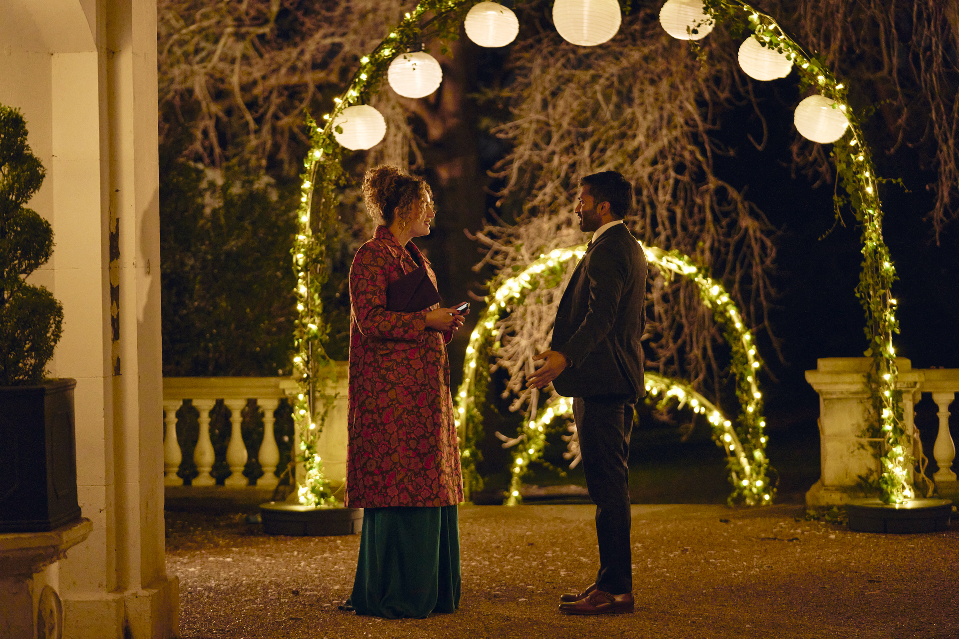 A man and a woman standing on a grand balcony lit with arches of fairy lights.