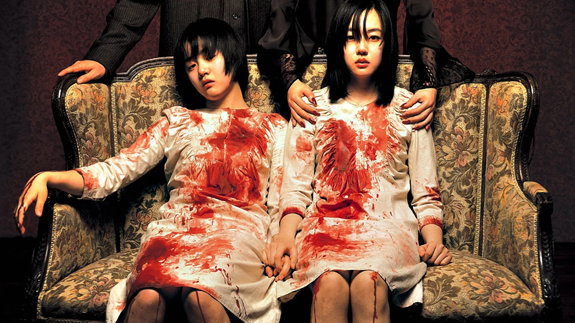 Two Korean women in white dresses covered in blood sit side by side on a brocade couch, one with an unseen figure standing behind and above her, hands firmly on her shoulders, in A Tale of Two Sisters