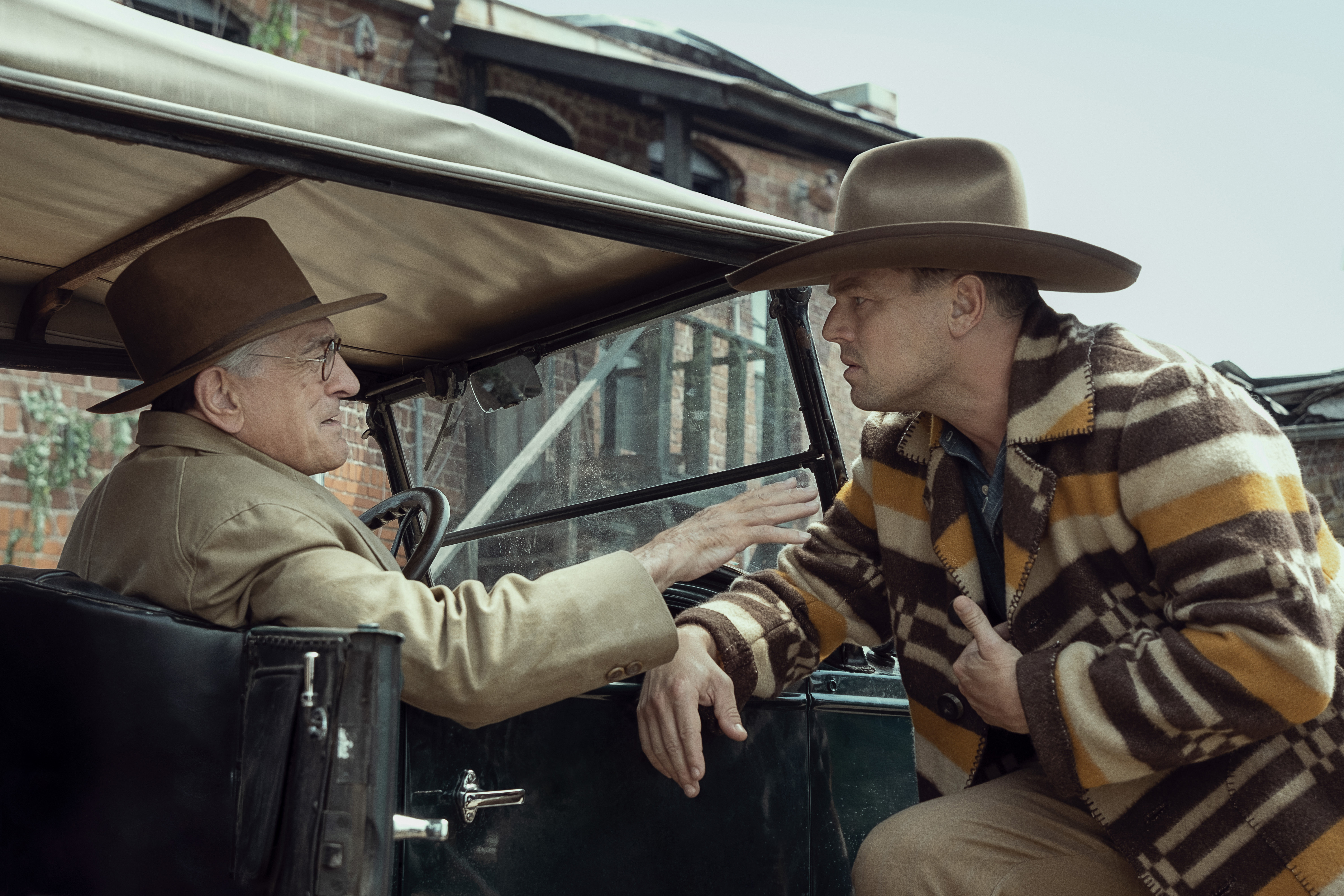 Leonardo DiCaprio in a cowboy hat and poncho leans over an automobile driven by Robert DeNiro in Killers of the Flower Moon.
