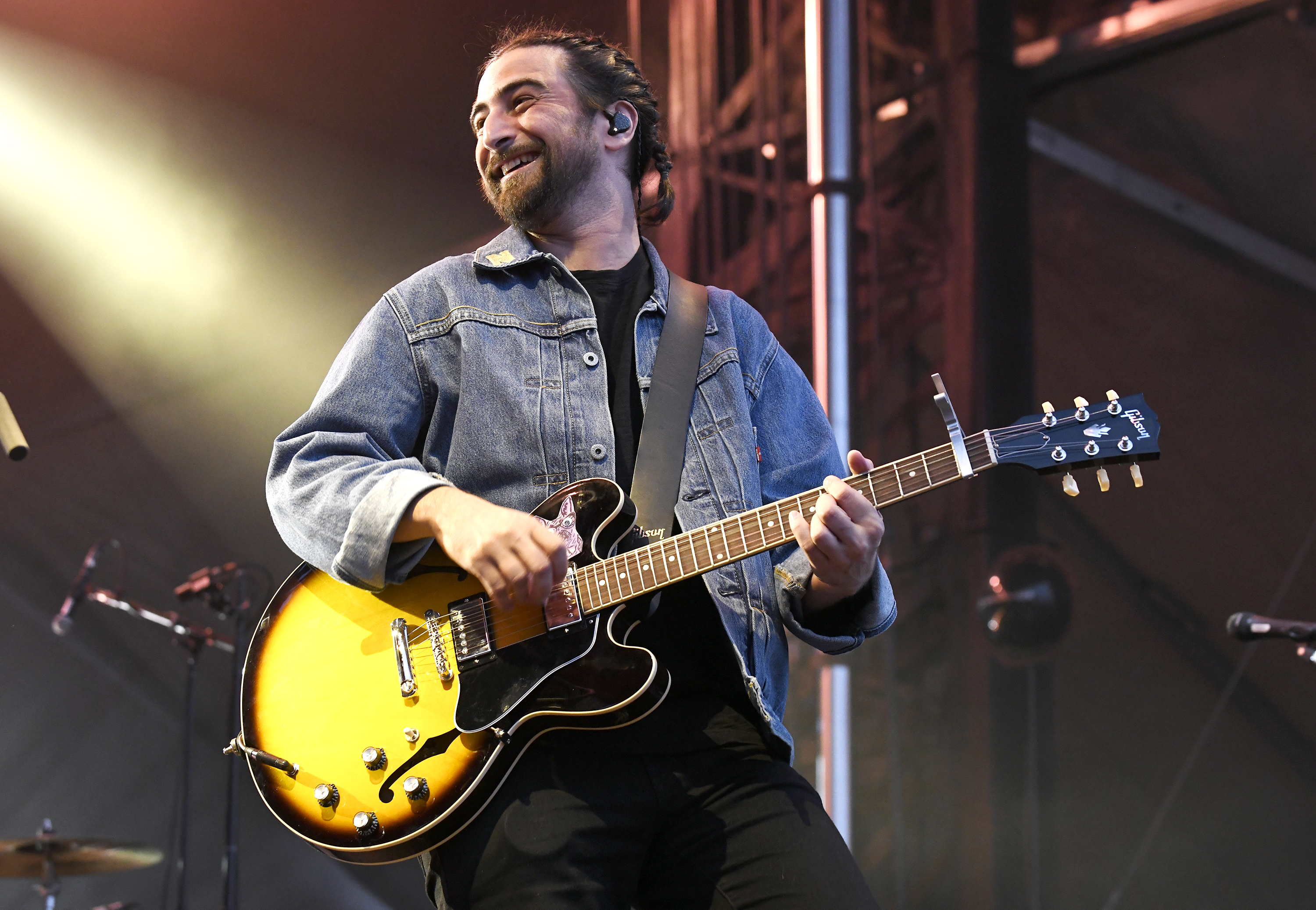 Kahan, a bearded man, with hair tied back in a braid, smiles while holding a guitar on stage.