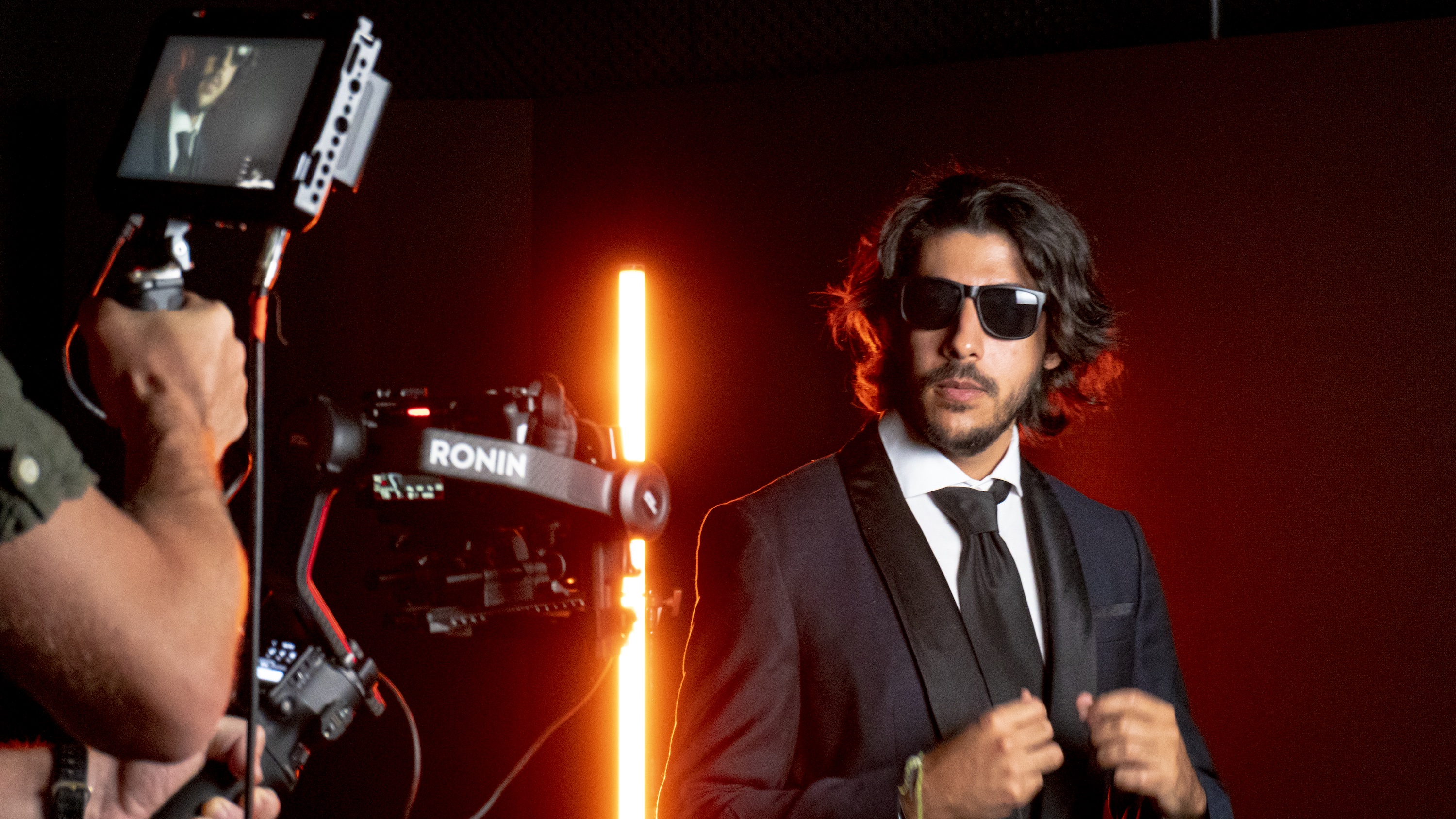Baiano poses for a video camera while wearing a black suit and black sunglasses