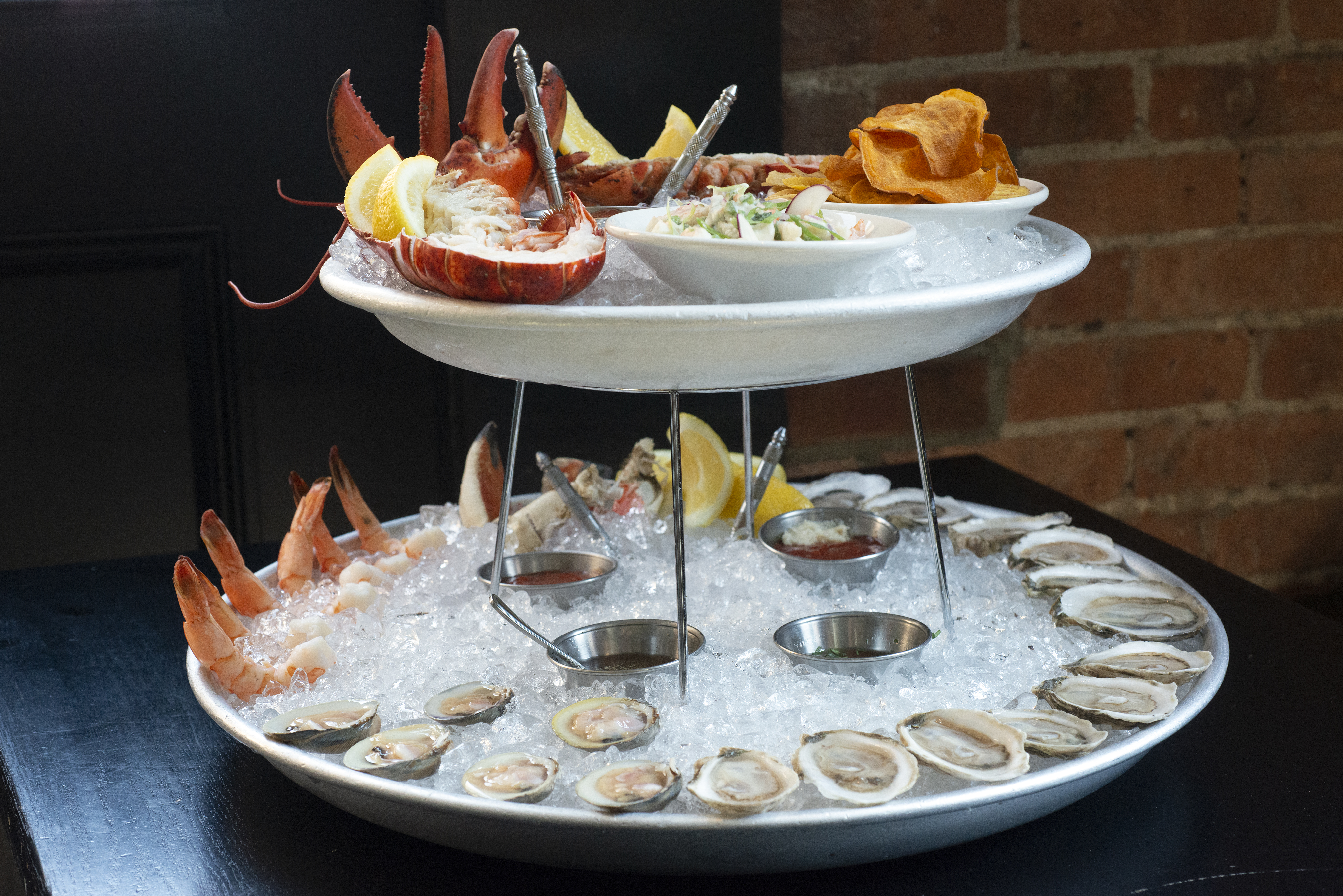 A two-tiered seafood tower with oysters, Jonah crab claws, shrimp, and more arranged around two steel platters filled with ice.