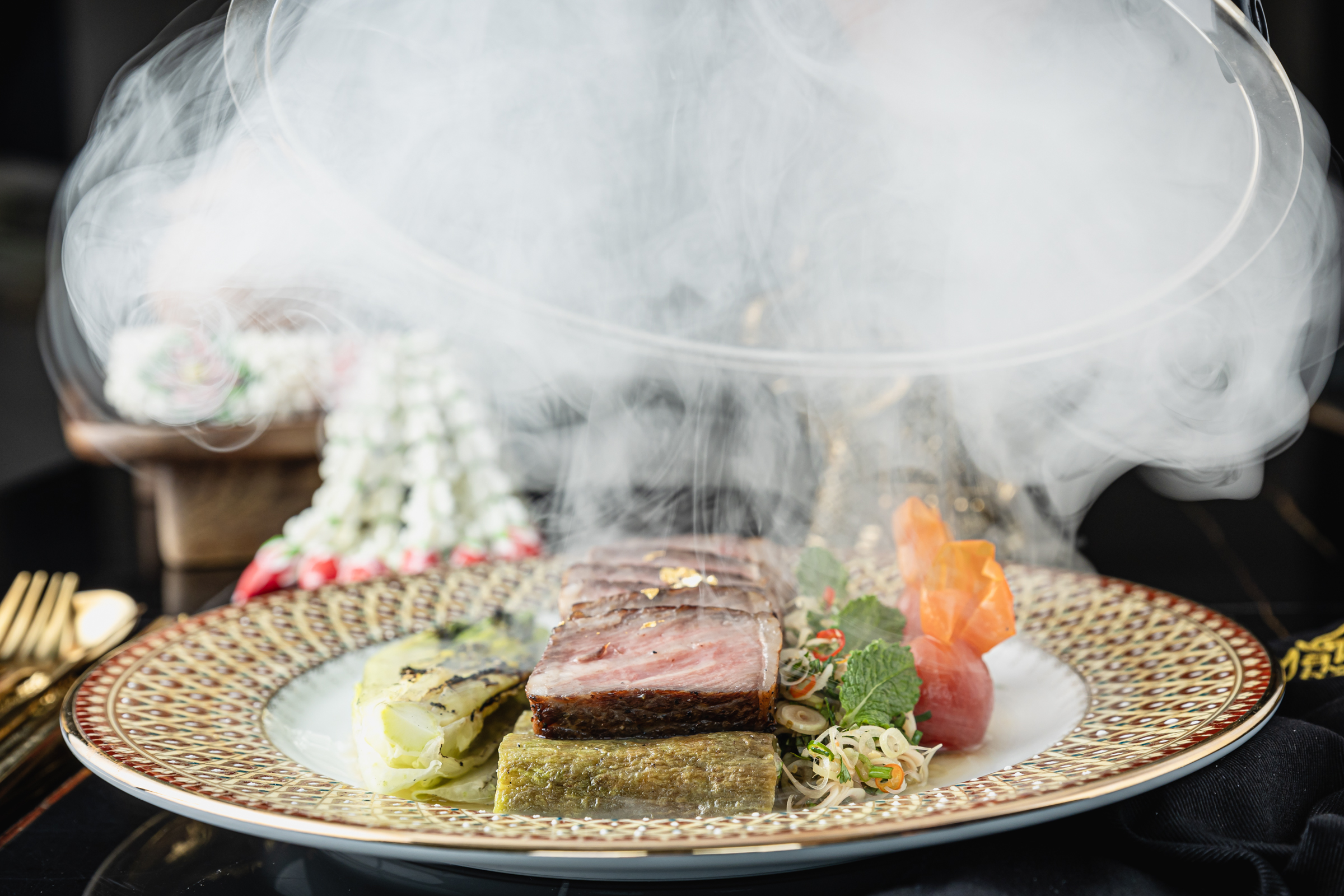 Slices of seared wagyu steak plated with charred eggplant, lemongrass, and carrots, with a cloud of smoke floating above.