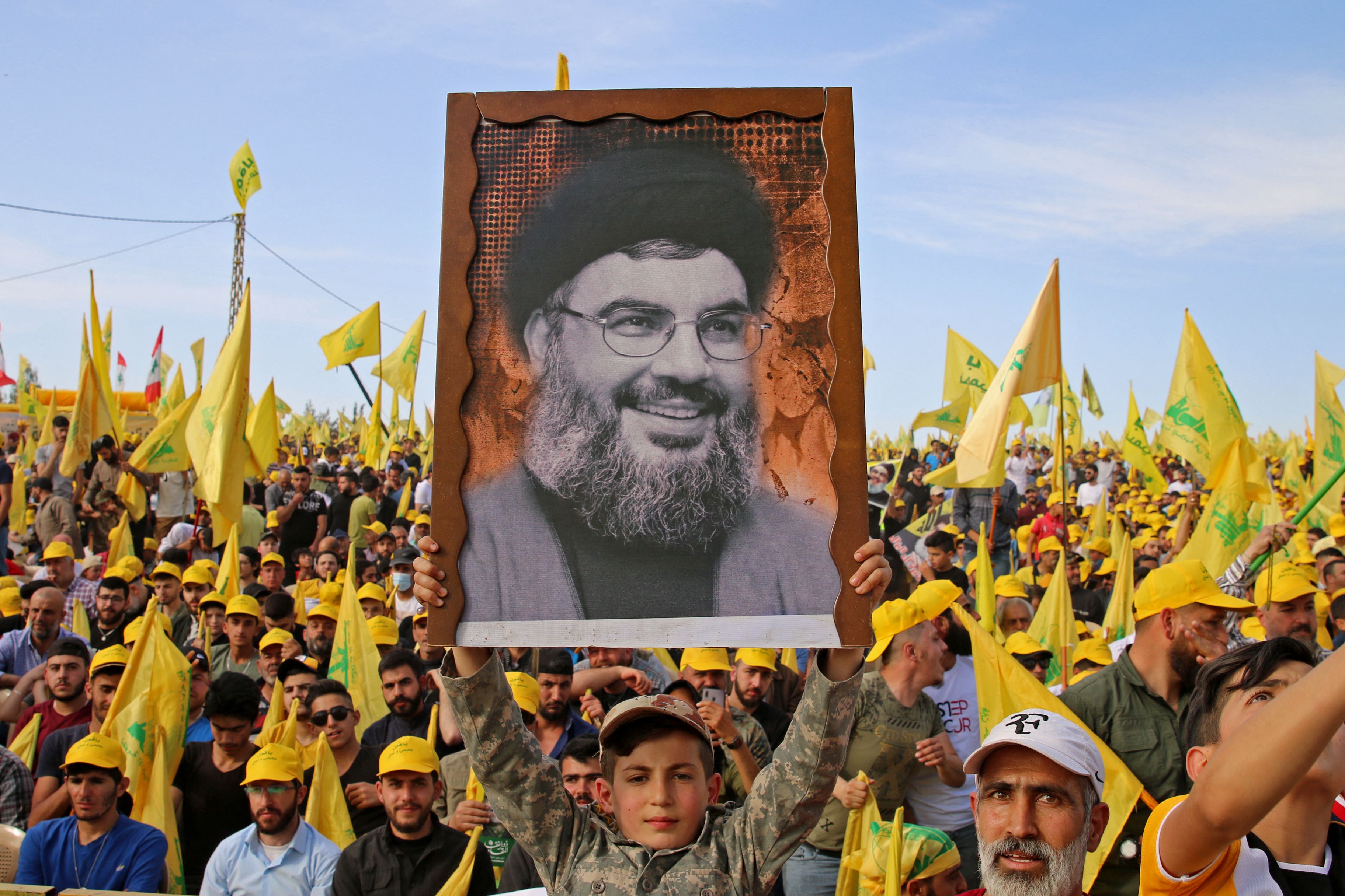 Bright yellow and green flags — Hezbollah’s colors — sprout from a sea of jubilant faces packed closely together. In the foreground, a young boy in fatigues raises a black-and-white photo of Nasrallah almost as big as he is above his head. In the image, Nasrallah, in glasses and a large beard, smiles in a fatherly way.