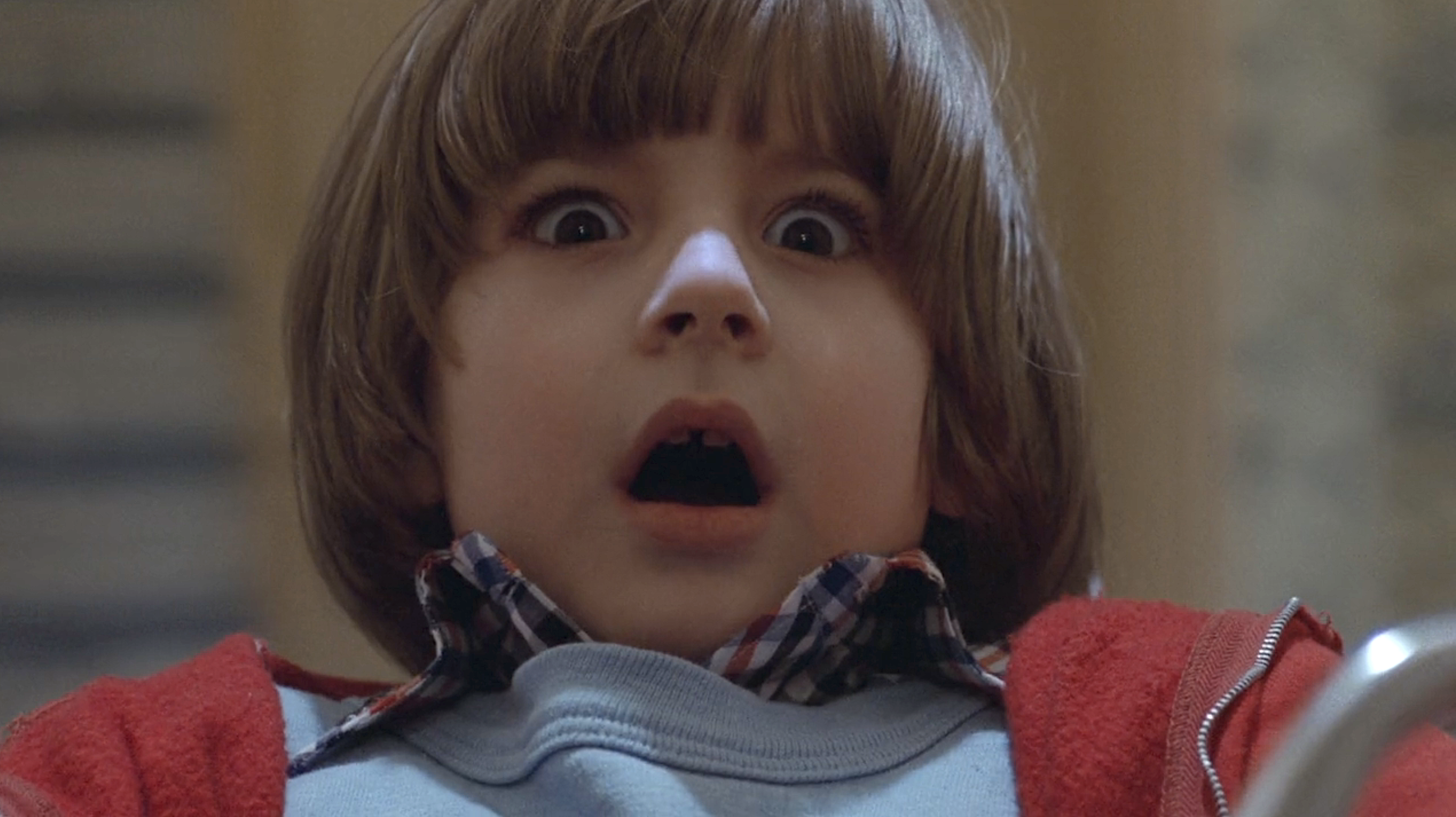 A closeup of Danny Torrance (Danny Lloyd), a very young child with bowl-cut hair and a red jacket, reacting with wide-eyed, mouth-open horror to the twin ghost girls in Stanley Kubrick’s movie adaptation of The Shining