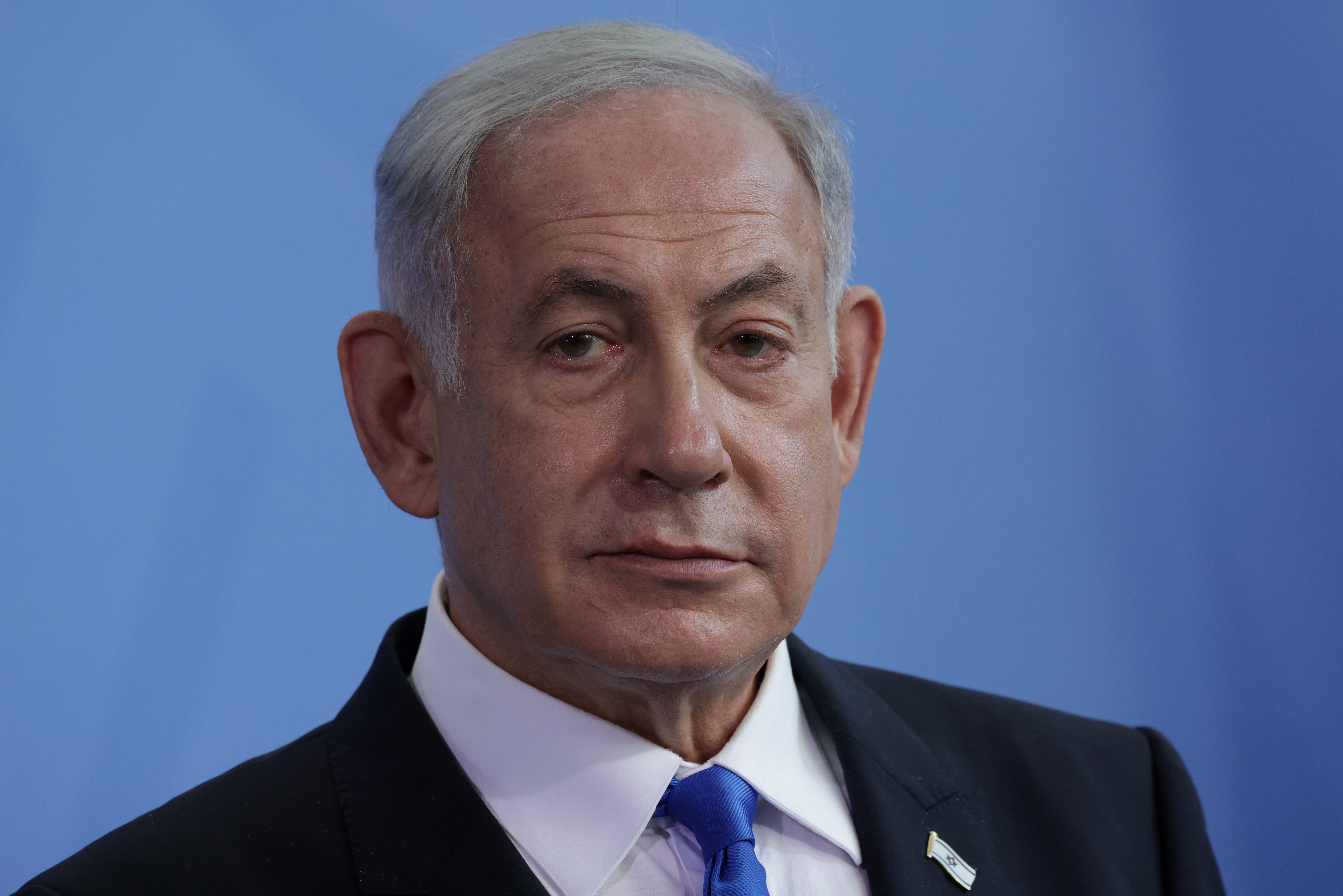Benjamin Netanyahu, in a black suit, white shirt, and blue tie, looks to the side during a media briefing in Berlin.