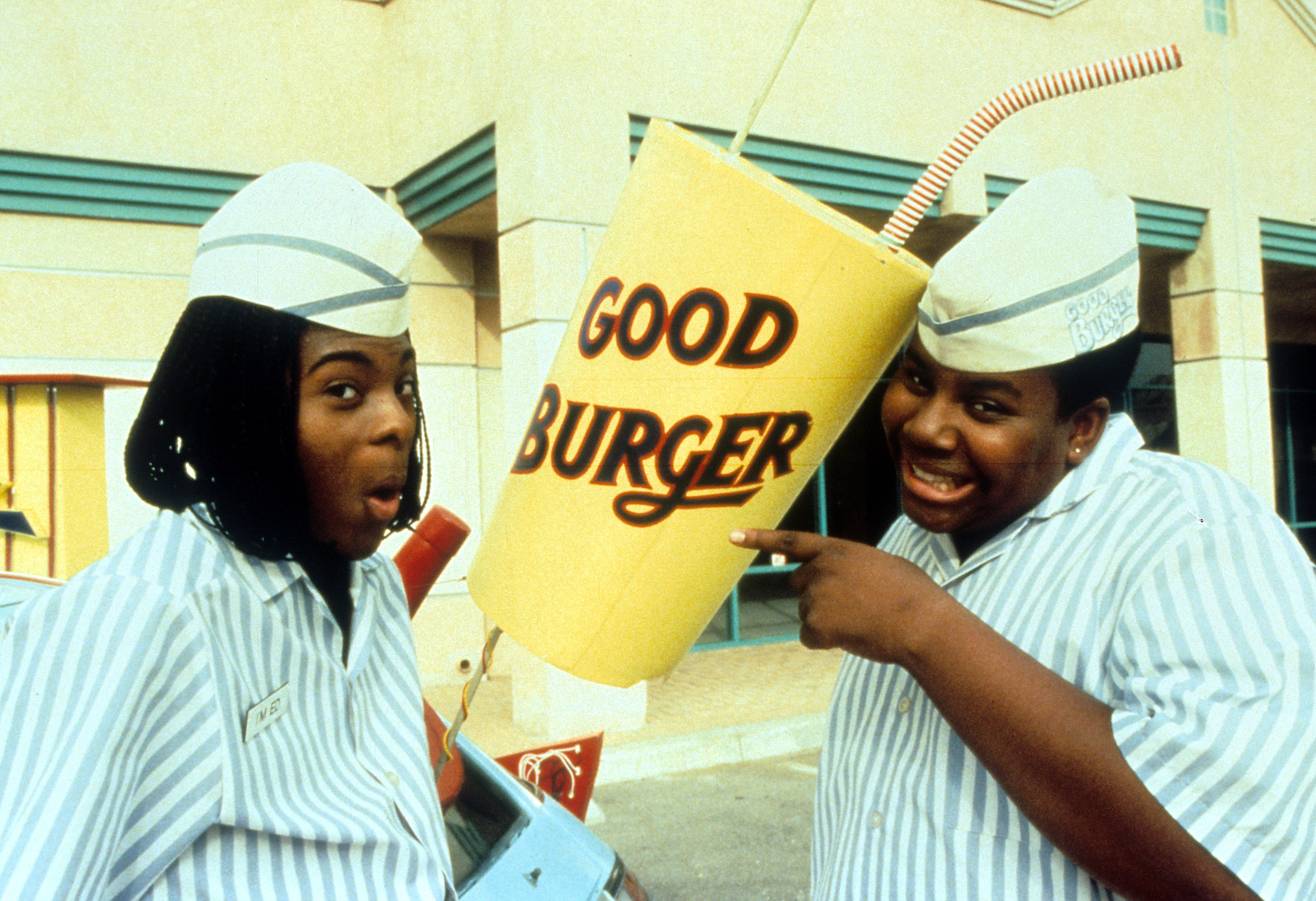 Two men dressed in fast food uniforms gesture to a giant soft drink cup with a “Good Burger” logo
