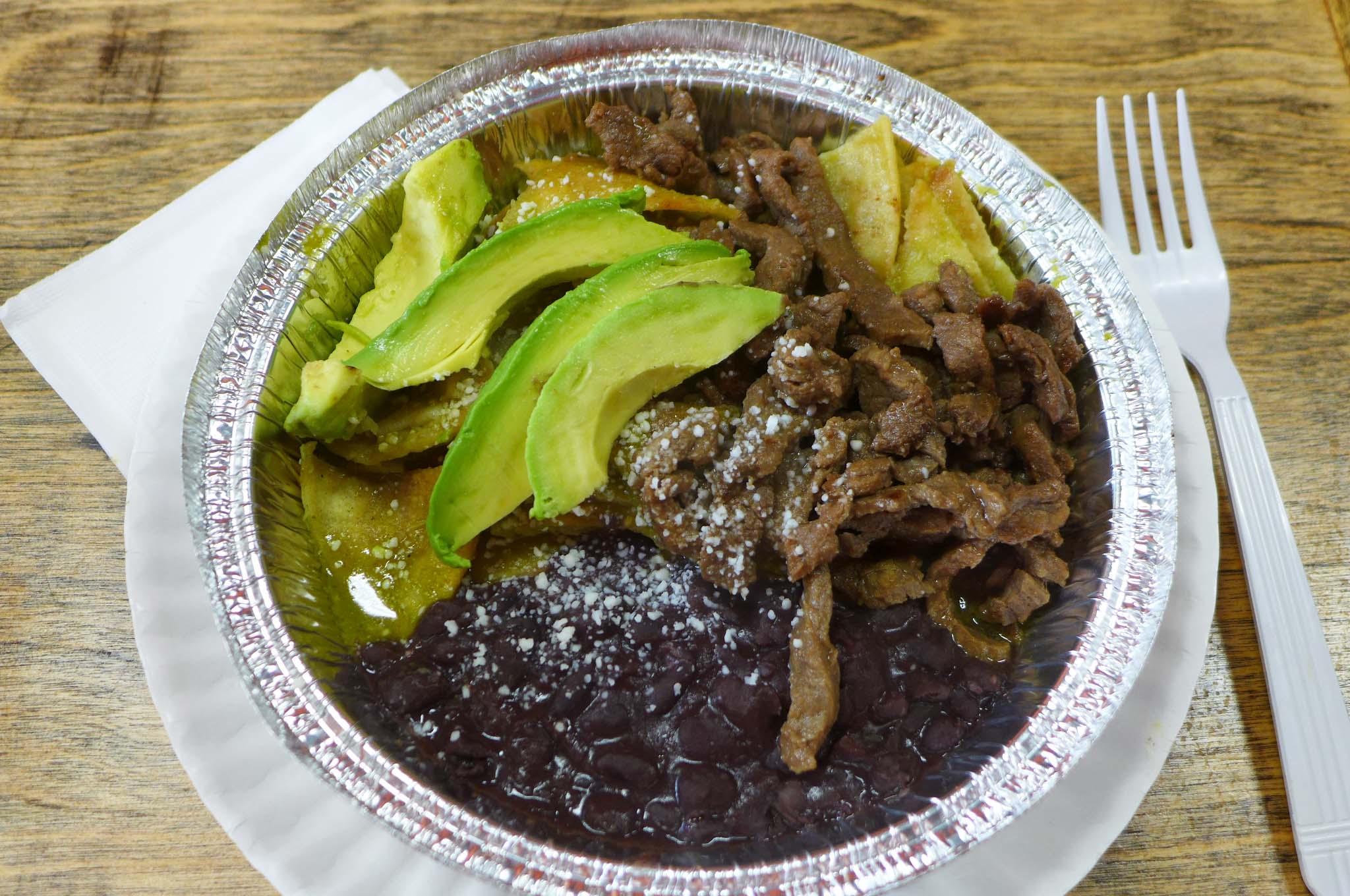 A round aluminum container with shreds of steak, beans, avocados and a few tortilla chips seen.