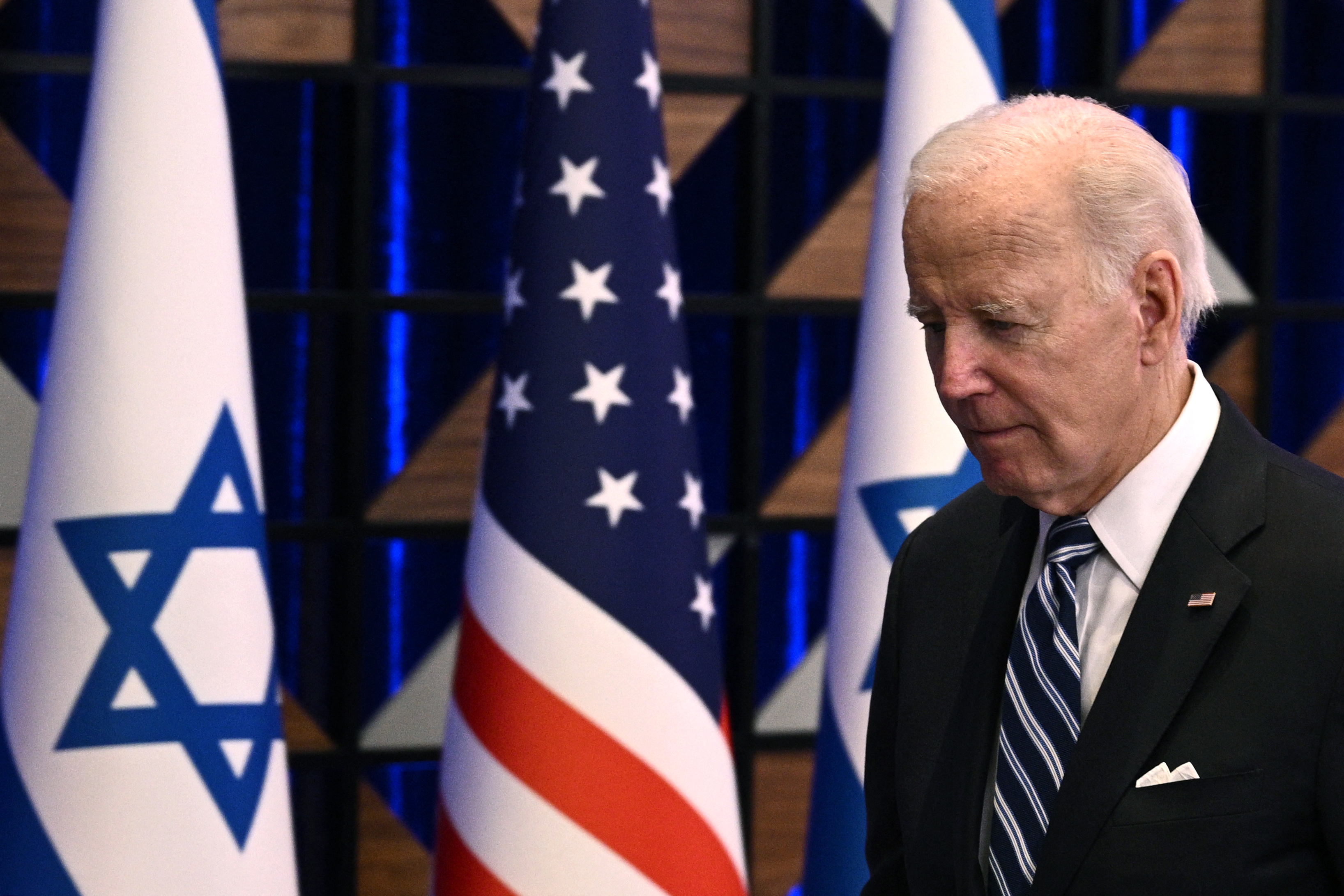 President Biden, in a dark suit and striped tie, looking slightly downward as he walks past the US and Israel flags on standing poles in a conference room.