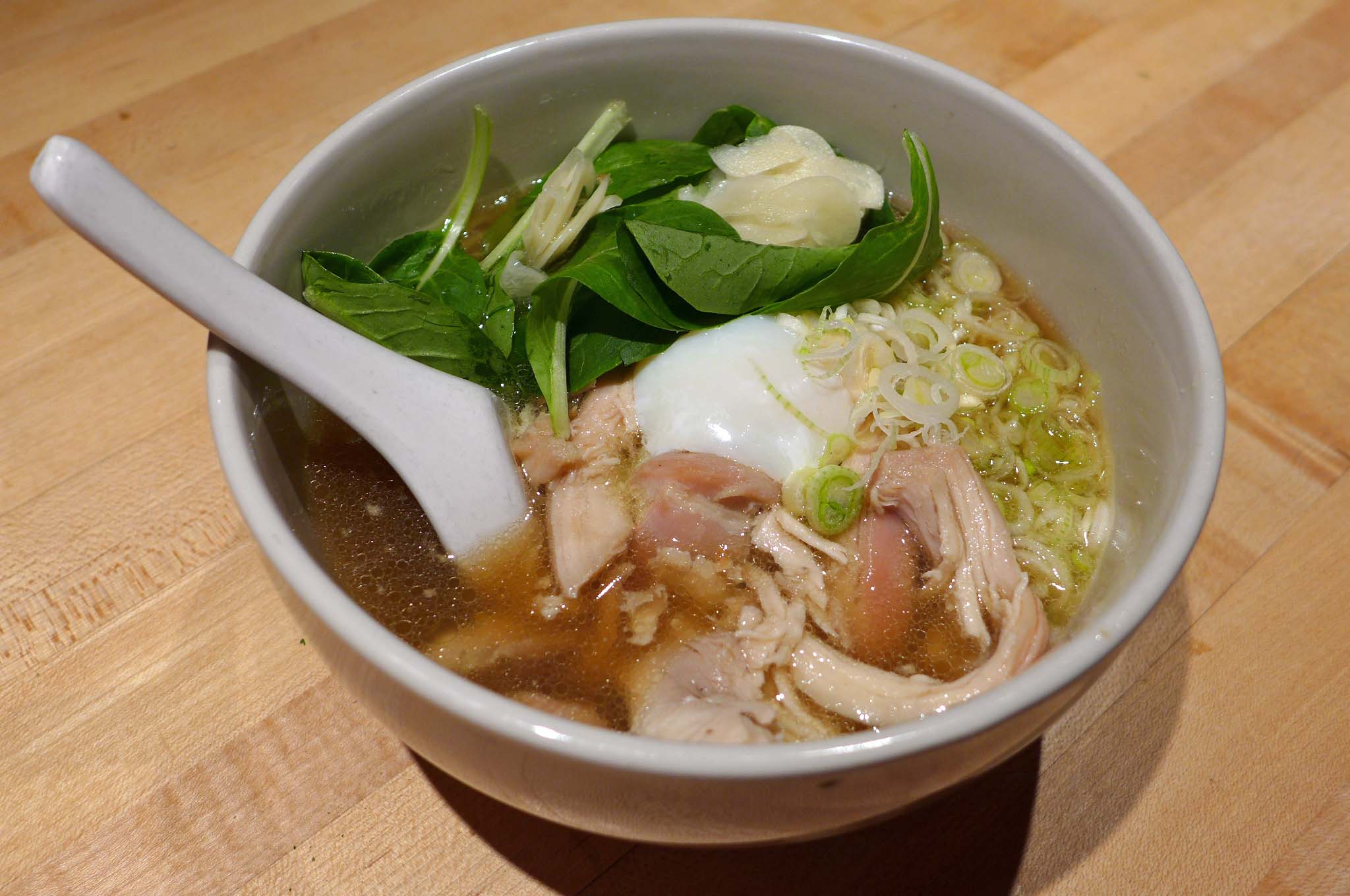 A bowl of medium brown broth with egg, raw spinach leaves, chicken, and noodles.