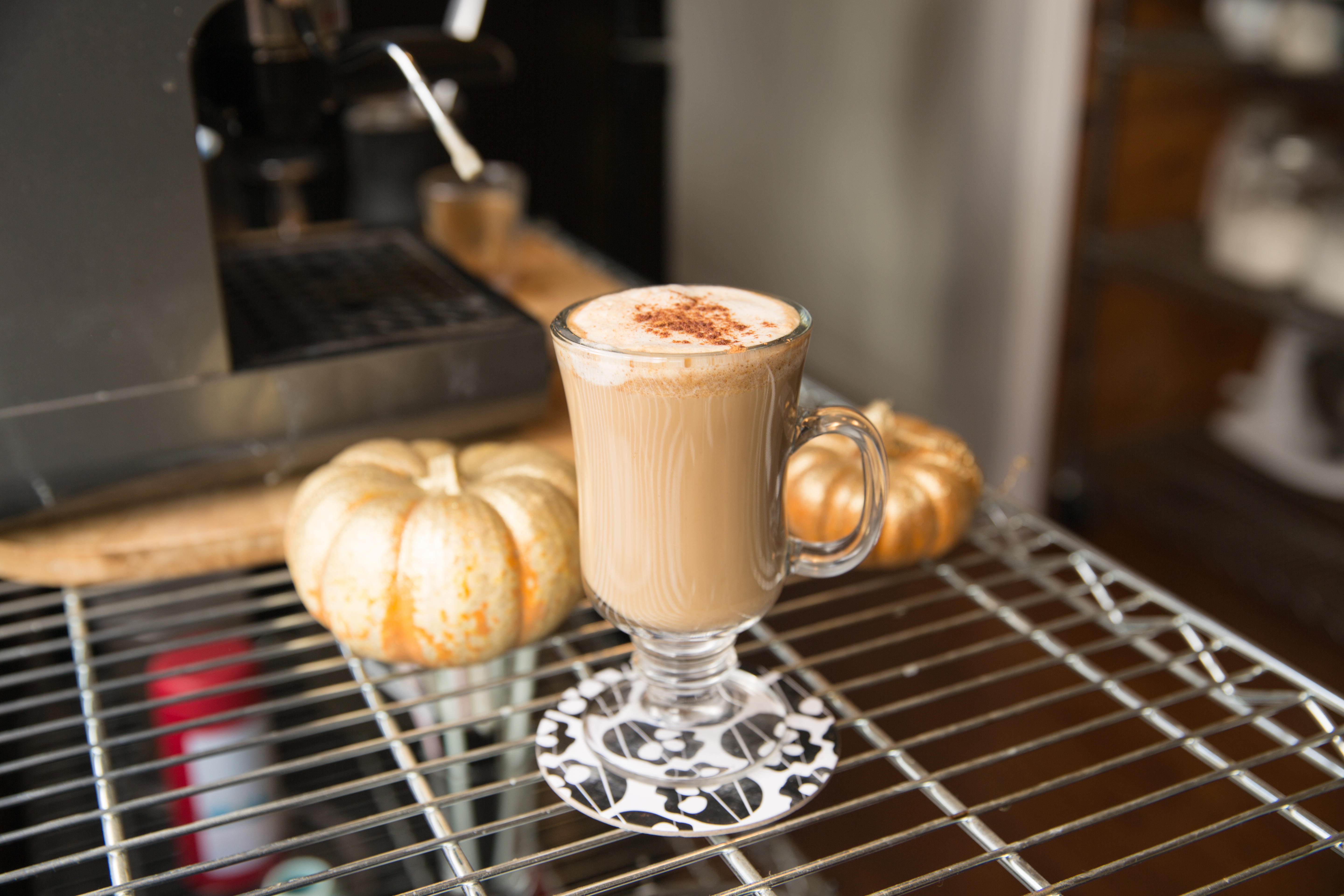 A freshly made pumpkin spice latte stands on a wire rack beside an espresso machine in a coffee shop.
