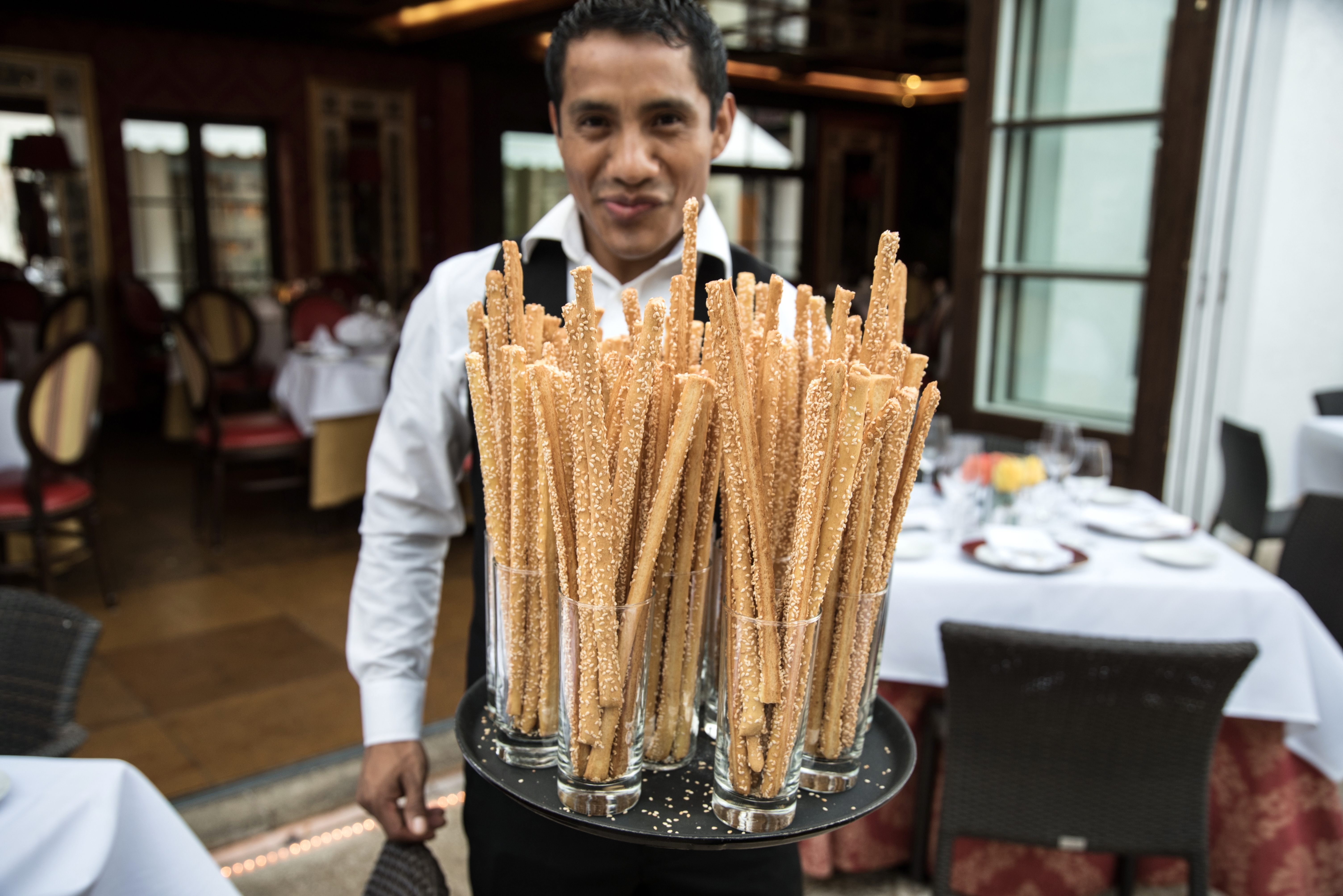 A waiter holds a tray full of grissini, Italian breadsticks. He’s looking directly at the camera. The courtyard of the restaurant is in the background and beyond the courtyard is the open doors of the inside of the restaurant.