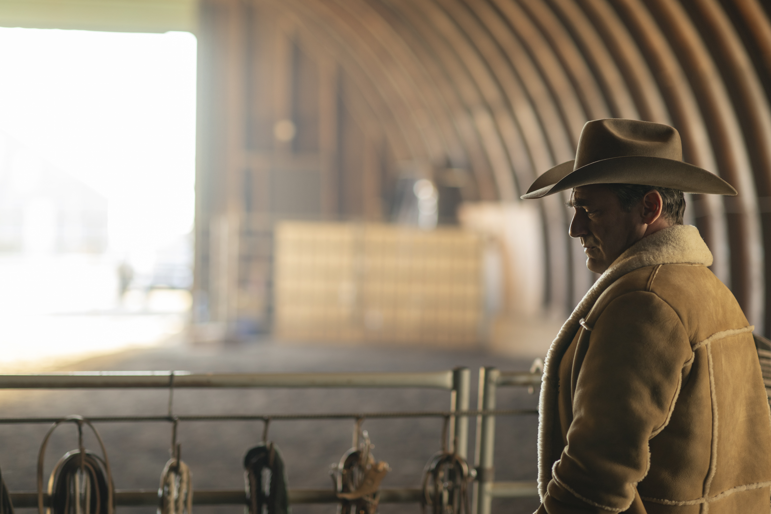Jon Hamm in a big shearling coat and cowboy hat looks thoughtfully over a giant barn in season 5 of Fargo