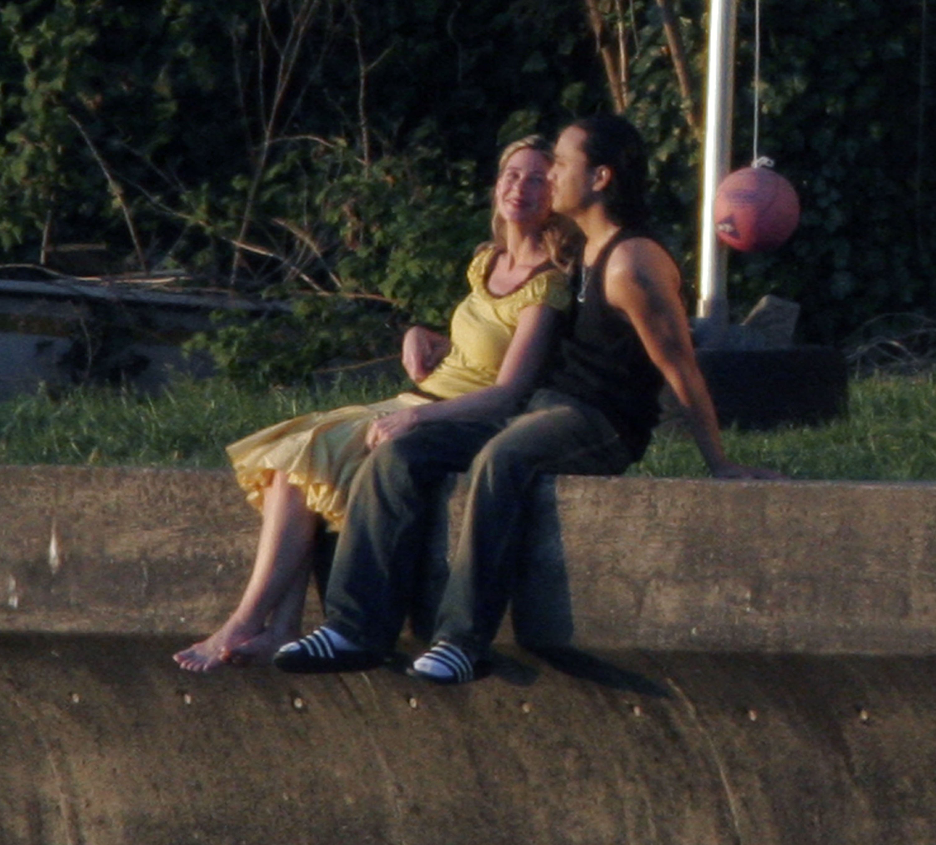 A couple sitting on a sunny concrete wall and leaning back on a grassy verge.