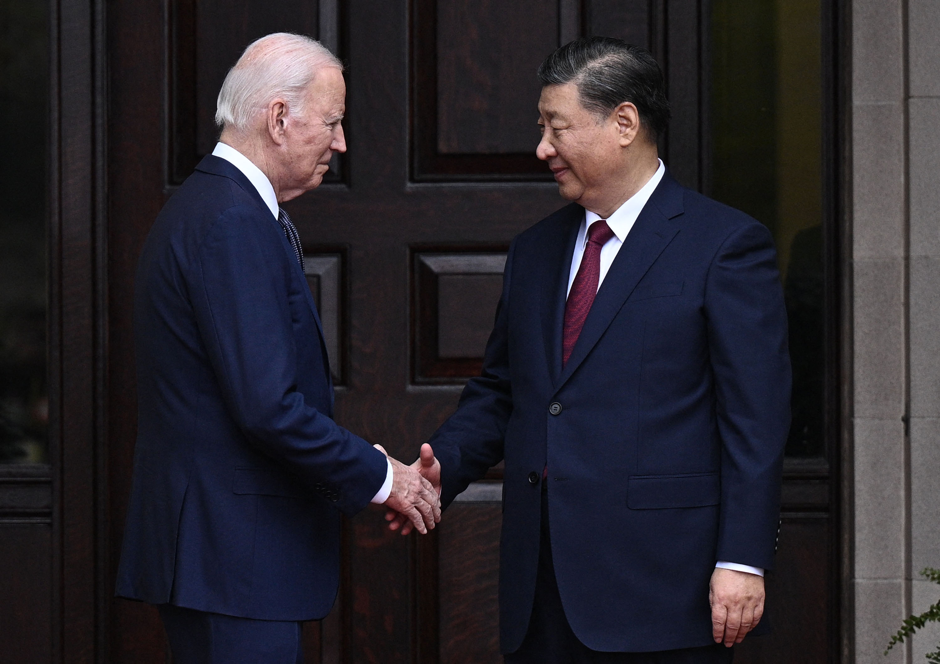 President Joe Biden, left, and Chinese leader Xi Jinping, right, shake hands while standing in front of a dark, paneled wood door.