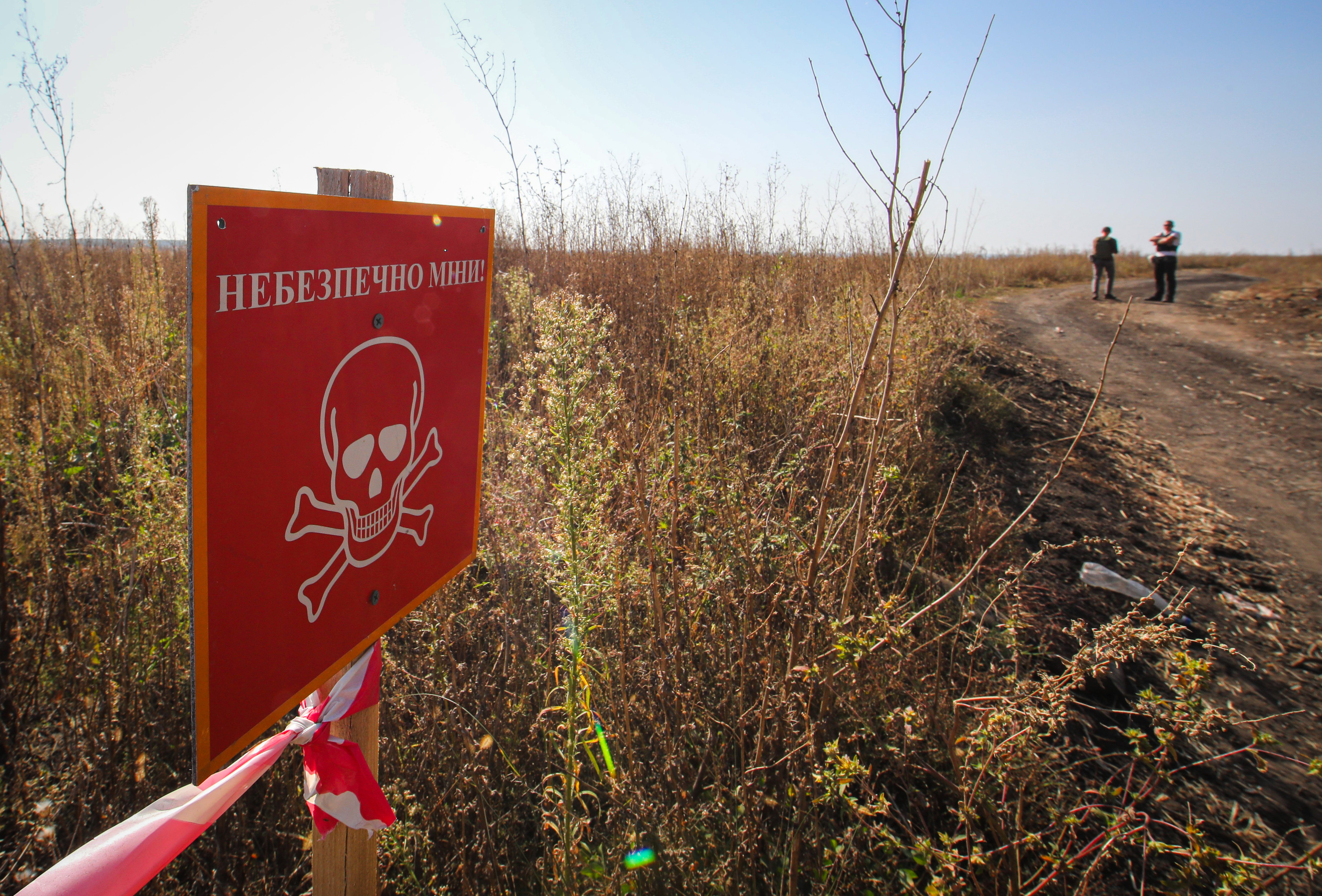 A red sign with a skull and crossbones reads, in Russian, “Danger. Mines.” It appears on a fence in front of an open field beside a dirt road.