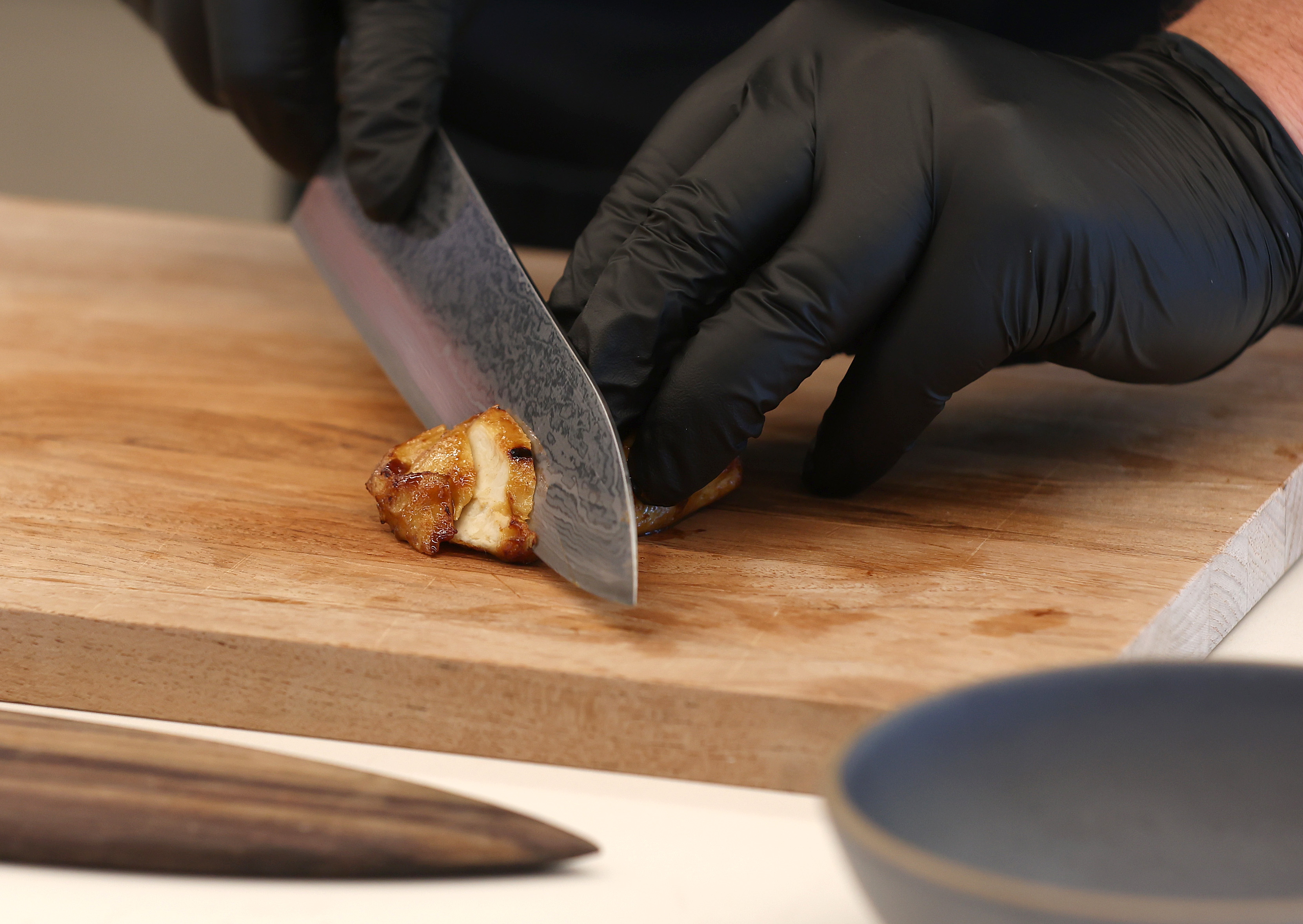Hands in black plastic gloves use a chef’s knife to cut a piece of cooked chicken on a board.