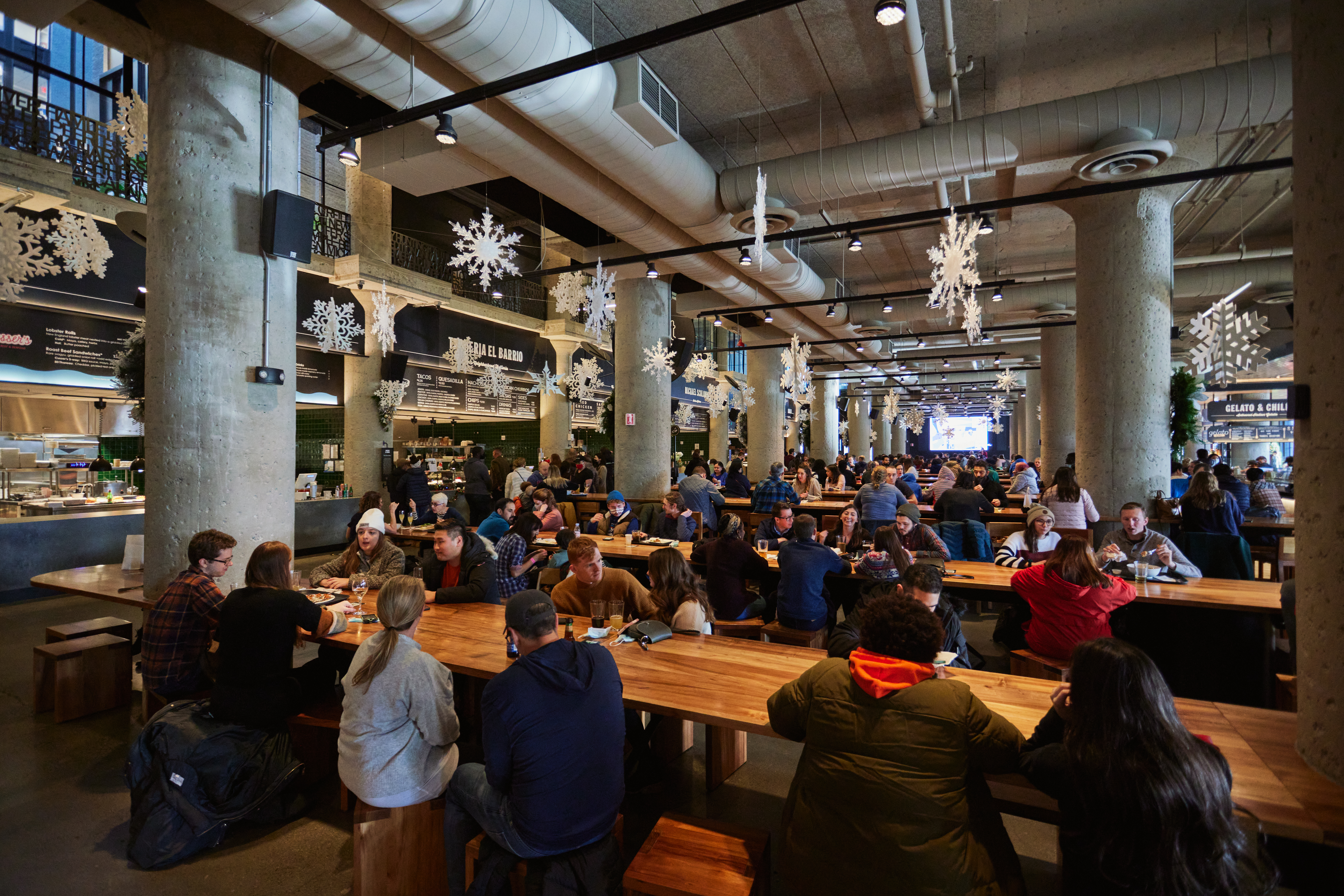 Snowflake decorations hang over long wooden tables where people eat and drink at a food hall.