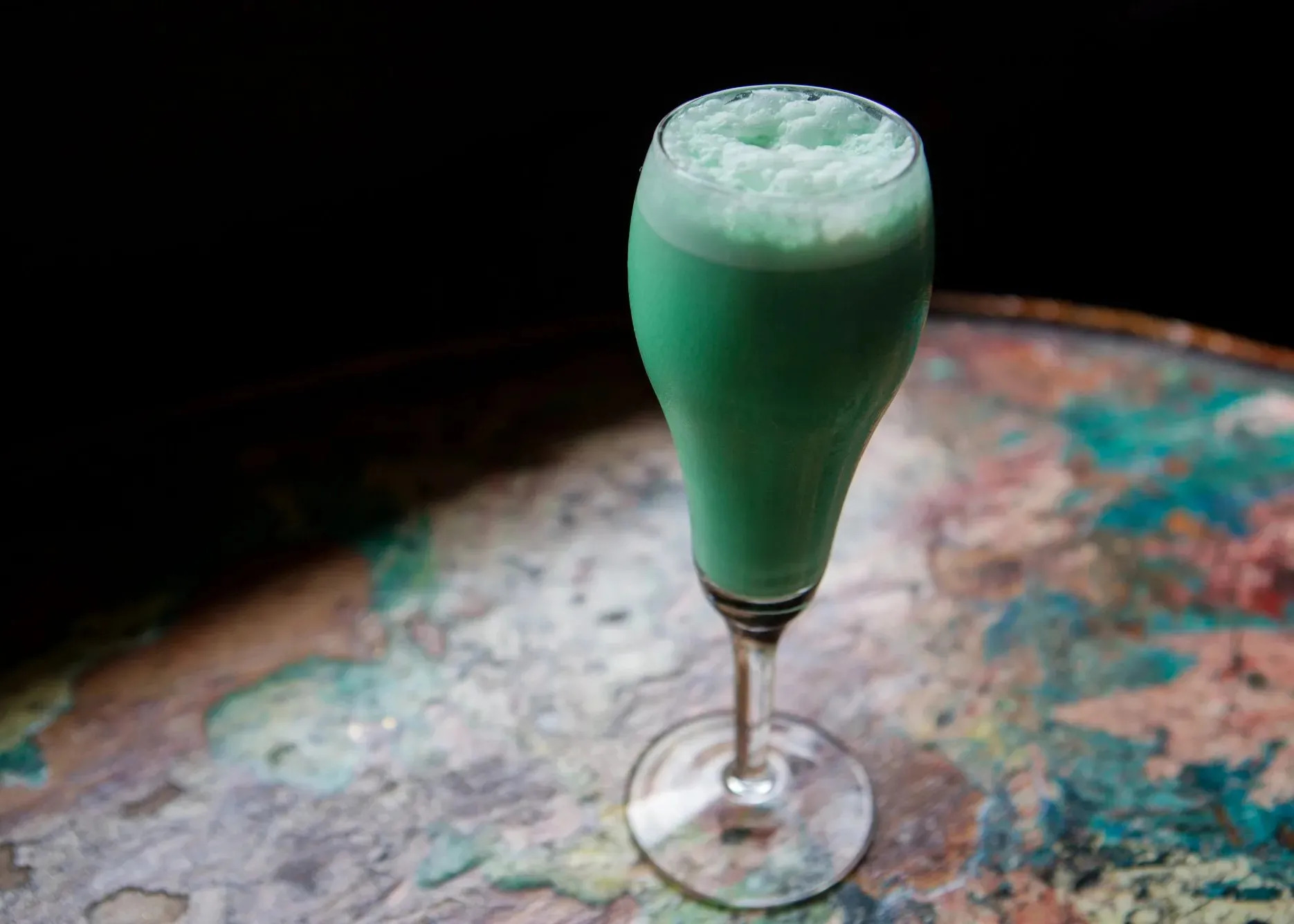 A bright green, frothy drink in a champagne-like flute sits on table.