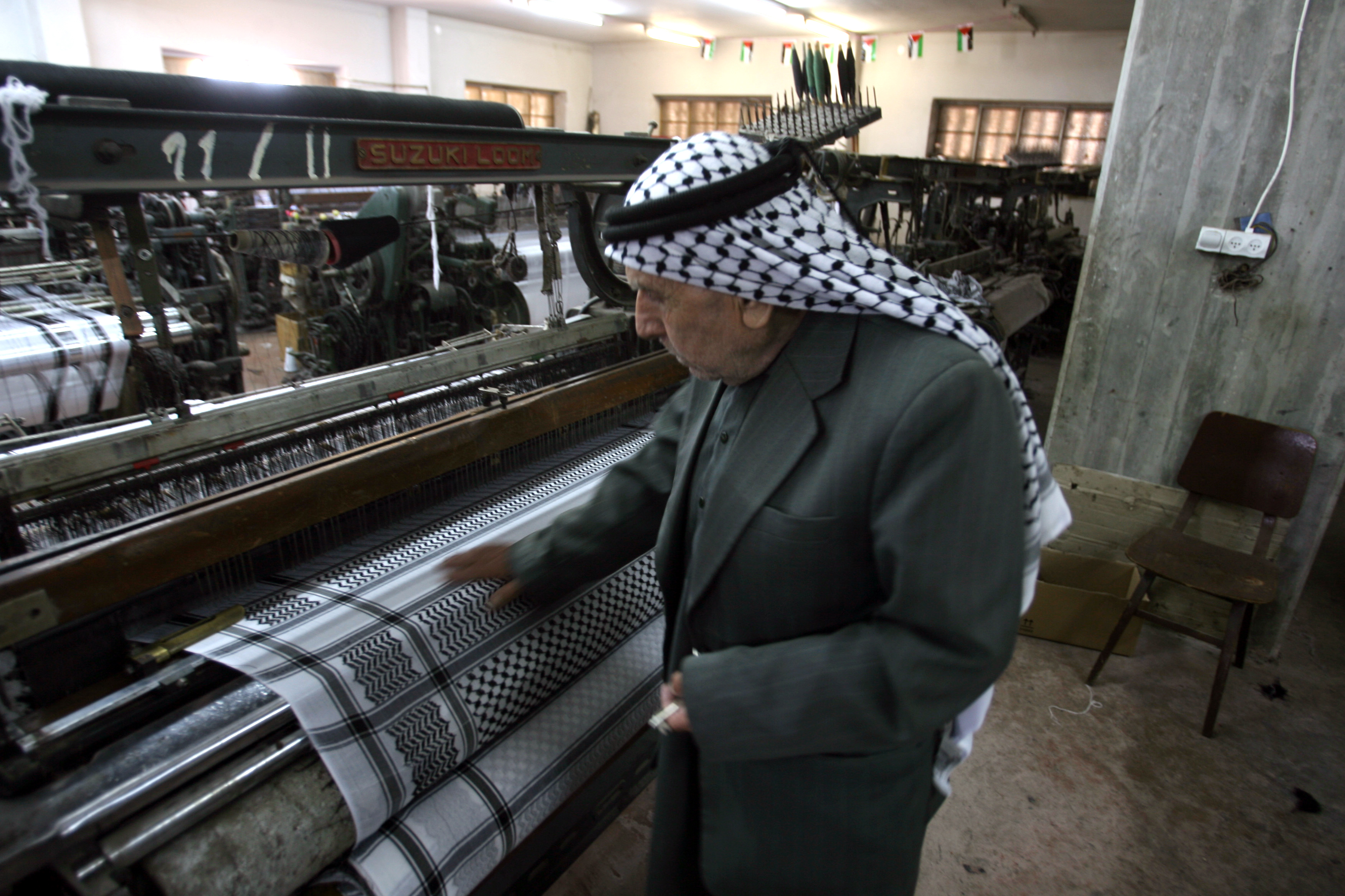 An elderly Palestinian man, Joudeh Hirbawi, is wearing a traditional keffiyeh on his head while standing by a machine at his factory as it sews a black-and-white keffiyeh, the iconic Palestinian scarf.