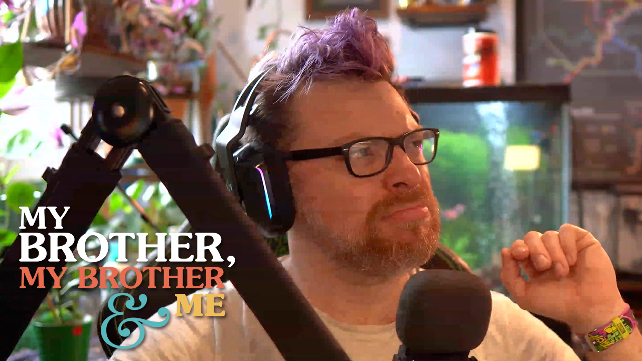 a photograph of Travis McElroy sitting in his office, behind a microphone. There are many plants and a fish tank behind him. He is wearing glasses and headphones, and he looks upset. The My Brother My Brother and Me logo is superimposed over the bottom left corner of the image.