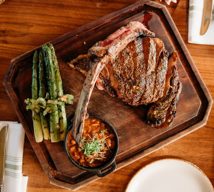 A steak with a bone on a board with green asparagus and a bowl of dark butter,