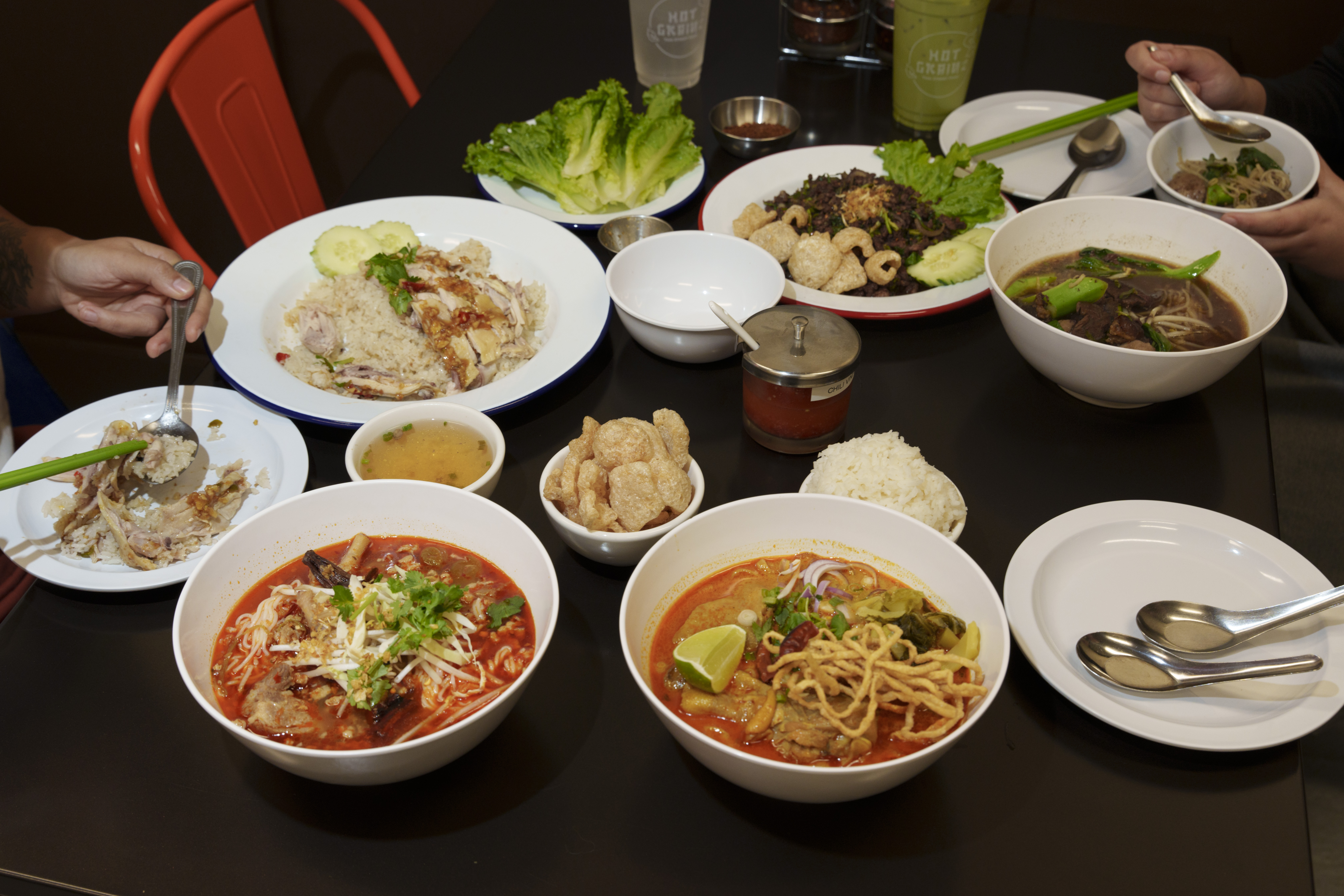 An assortment of white bowls and plates with various Thai foods on a black table, and two sets of hands holding utensils visible on either side.