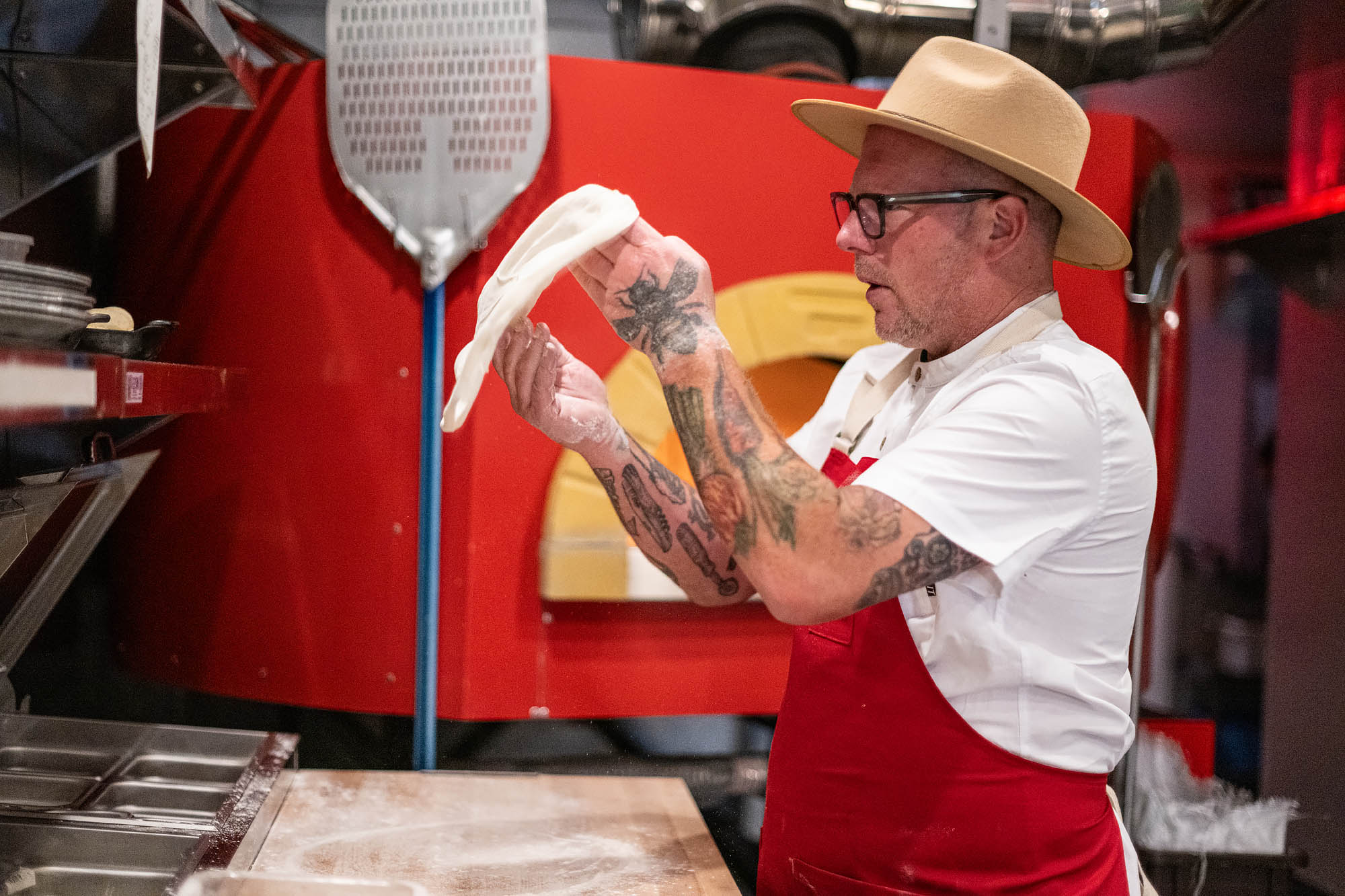 Jason Neroni, wearing glasses and a hat, stretches pizza dough at his new restaurant Best Bet in Culver City with a red pizza oven behind him.