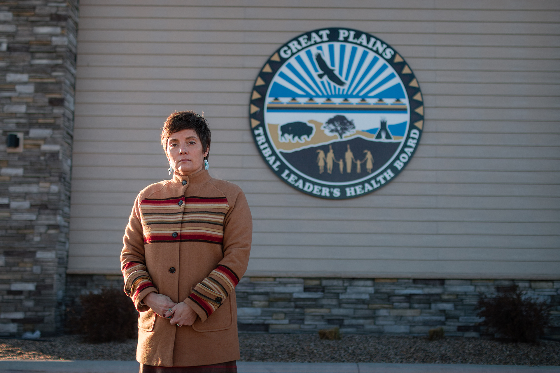 A woman with short hair stands in front of a building with stone details. A large circular sign reads “Great Plains Tribal Leader’s Health Board.”