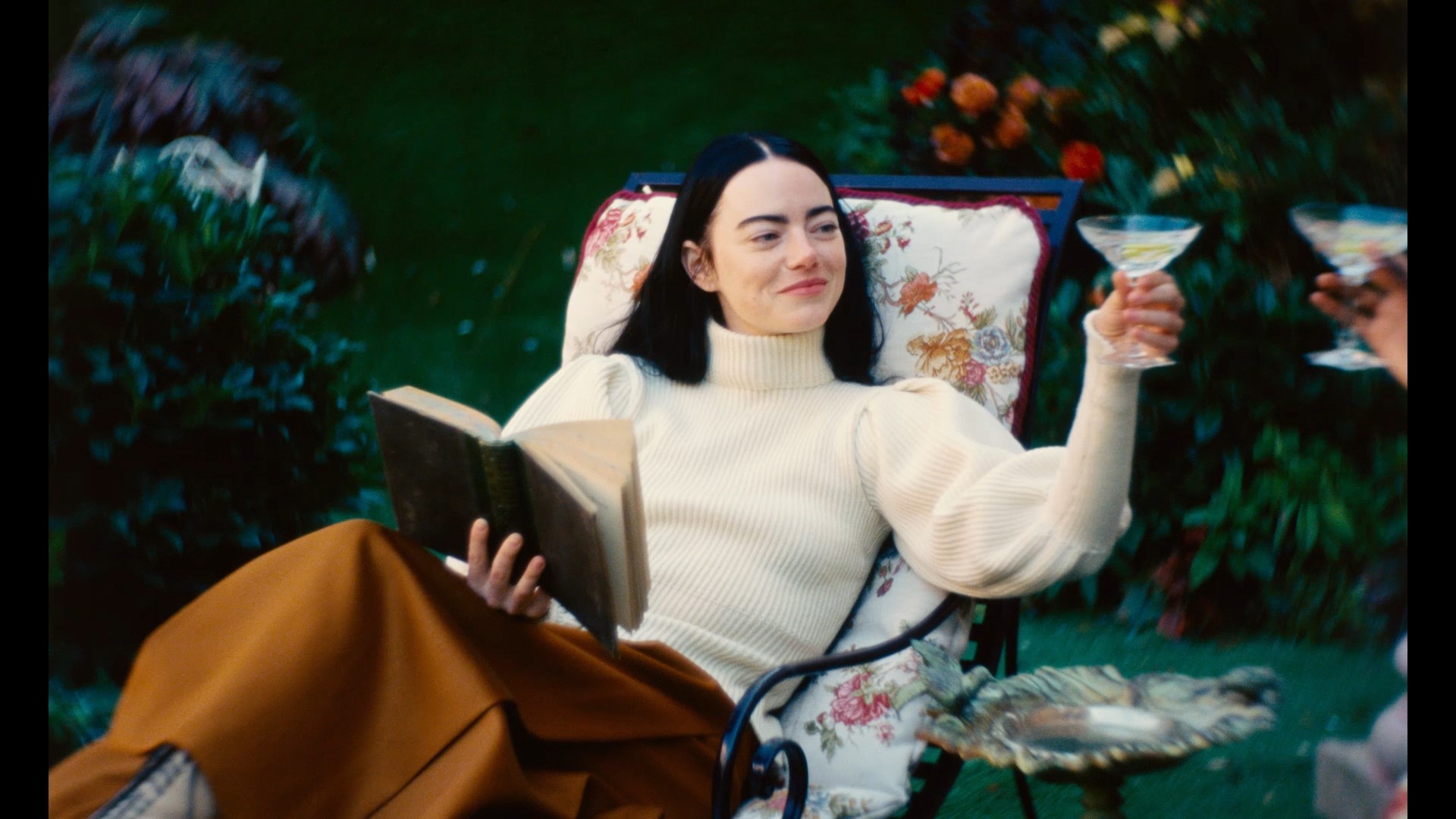 Emma Stone as Bella Baxter lounging in a garden chair, holding a book, smiling, and raising a cocktail glass in Poor Things