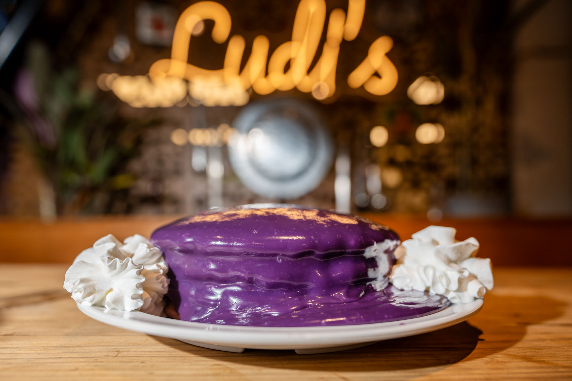 A plate of pancakes covered in bright purple ube sauce under a neon sign that says “Ludi’s.”