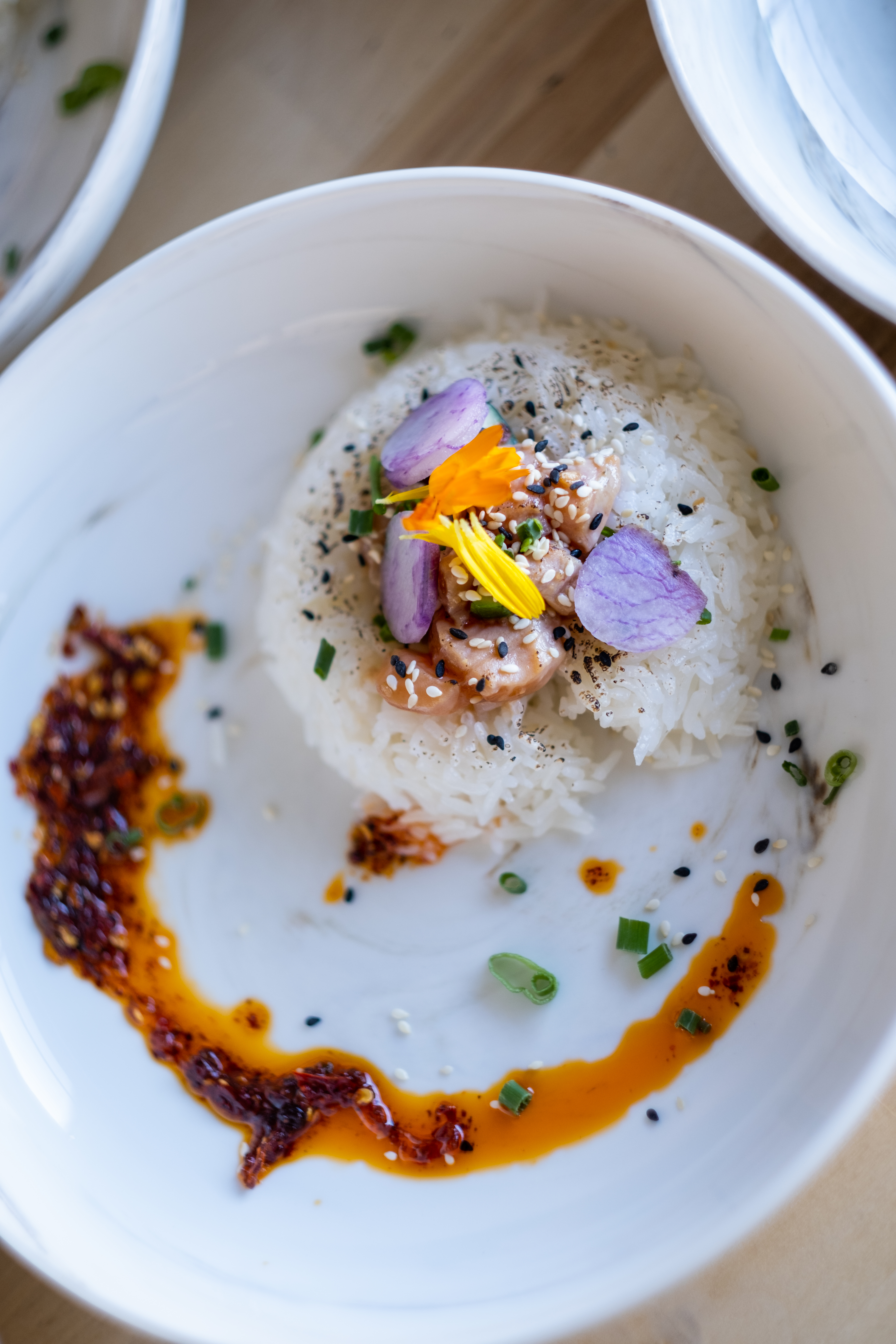 A white dish with a mound of rice topped with flowers, black sesame seeds, and salmon, with a crescent of chili oil on the plate. 